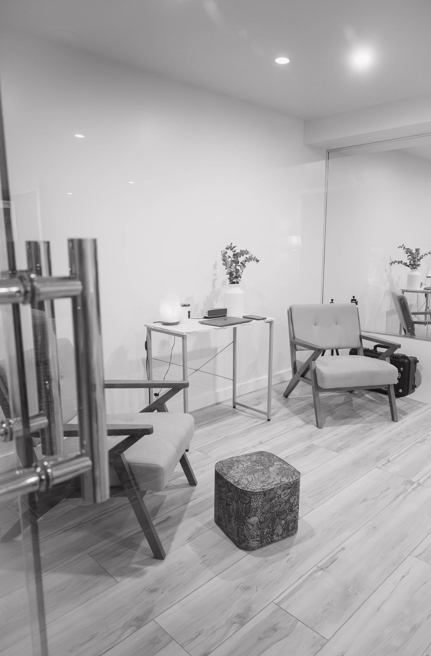 Black and white photo of a modern minimalist sitting area with wooden chairs, a side table with vase, and light hardwood floors.
