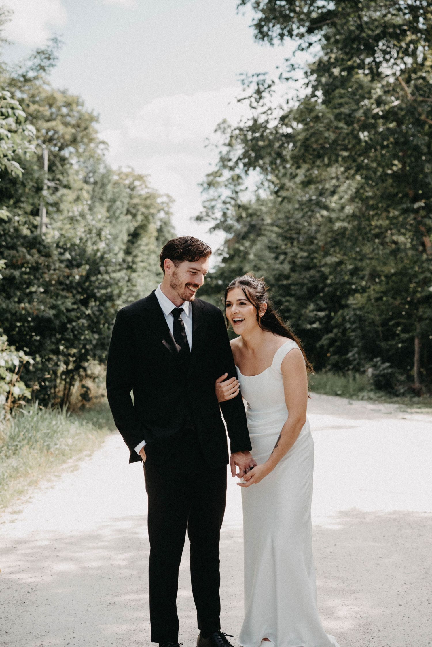 A smiling couple in wedding attire walks hand-in-hand on a tree-lined path, bride in white gown and groom in black suit.