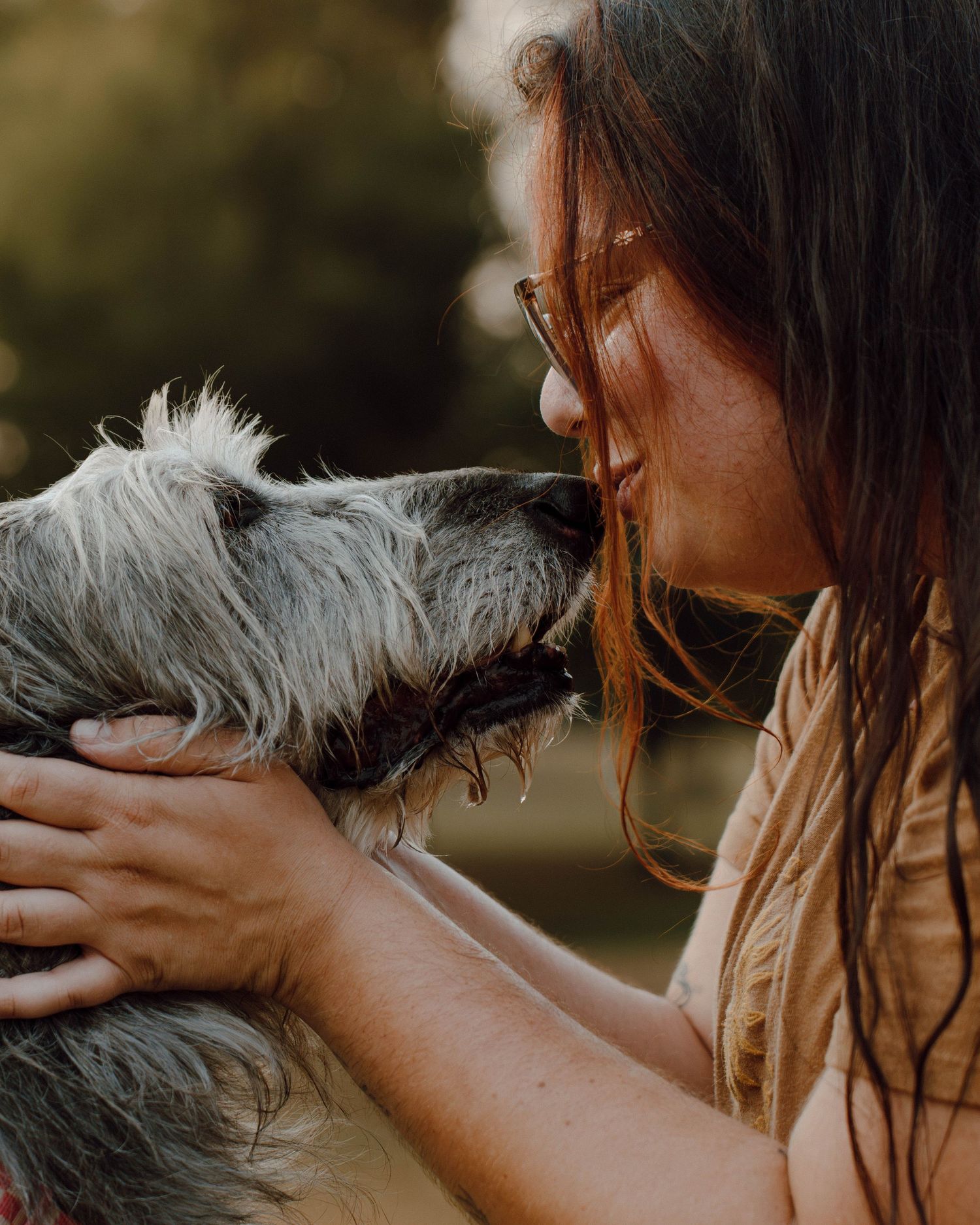A young woman with brown hair shares a tender moment kissing her scruffy gray dog outdoors.