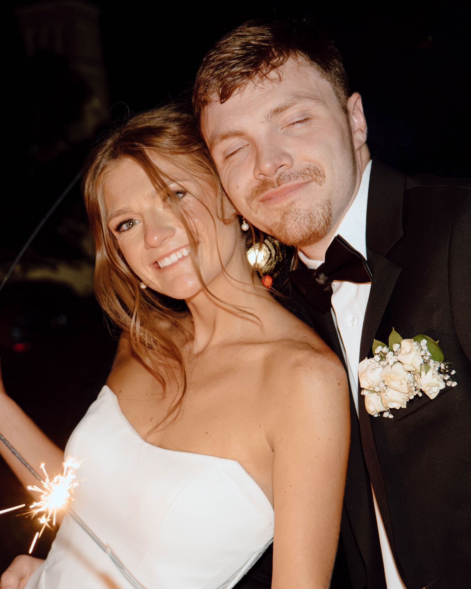 A smiling bride in a white strapless gown holds a sparkler beside a groom in a tuxedo with a floral boutonniere.