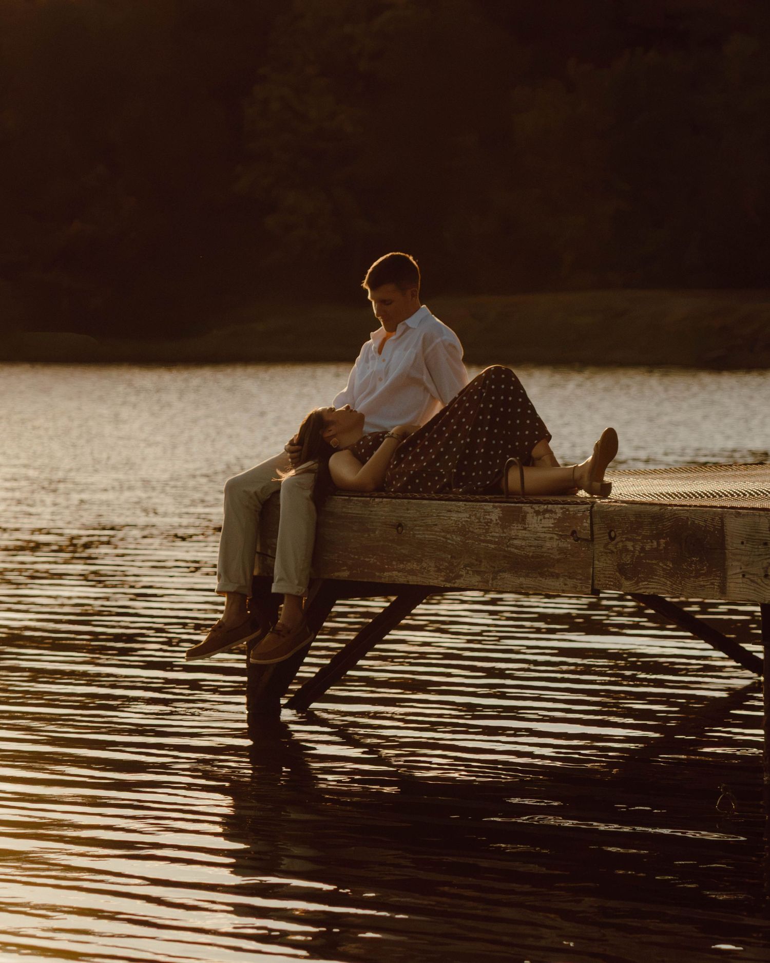 Couple relaxing on a wooden dock at golden hour, with one person lying in the other's lap by a calm lake.