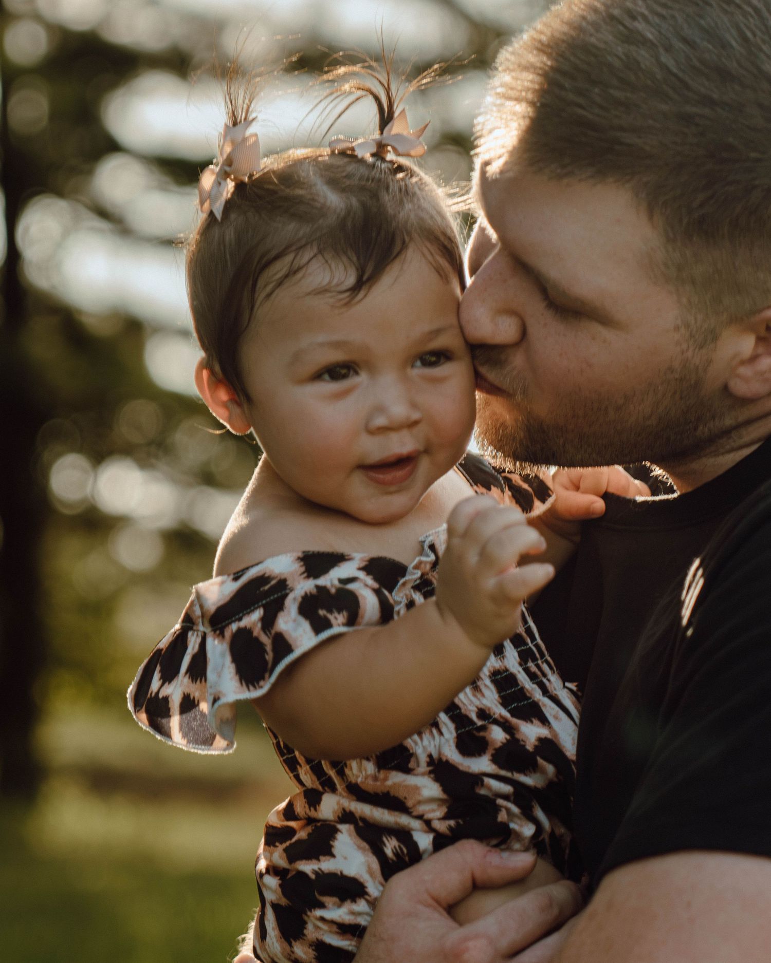 Father kissing baby girl in leopard print outfit outdoors with soft bokeh background.