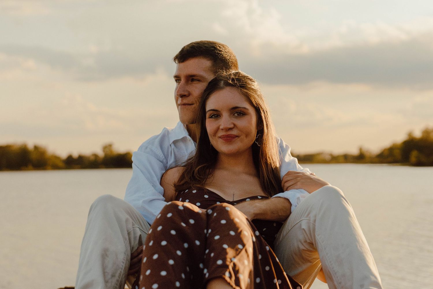 Couple sitting together by a lake at sunset, woman leaning back against man, smiling warmly in golden light.