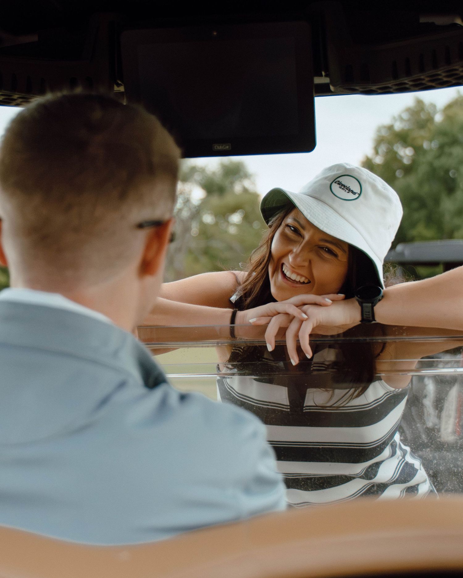 Smiling woman in bucket hat leaning on golf cart roof looking at man inside.