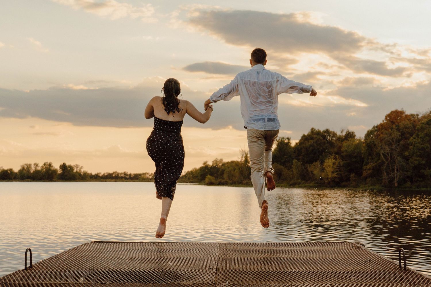 Couple holding hands jumping off a dock into a lake at sunset with trees in the background.