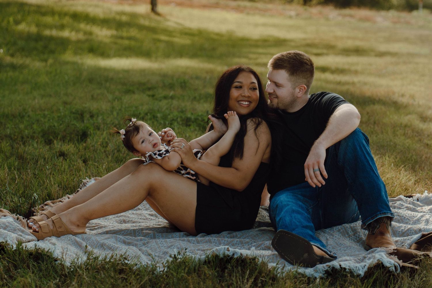 Family sitting on blanket in park, parents smiling at each other with baby girl in leopard print outfit.