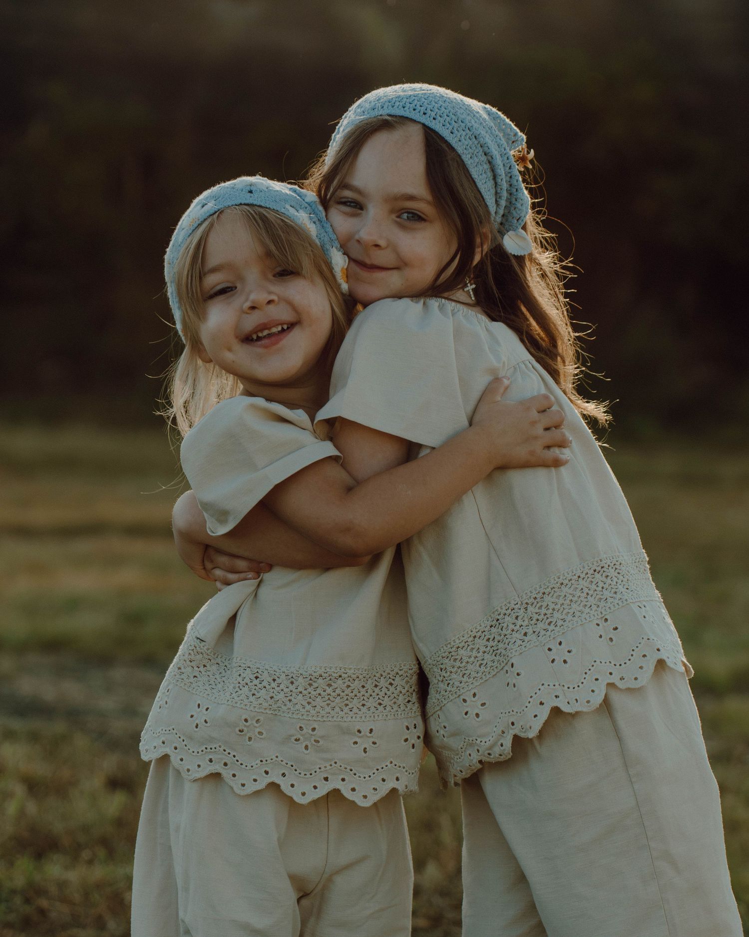 Two smiling young sisters in matching cream dresses and blue knit headbands hugging outdoors at golden hour.
