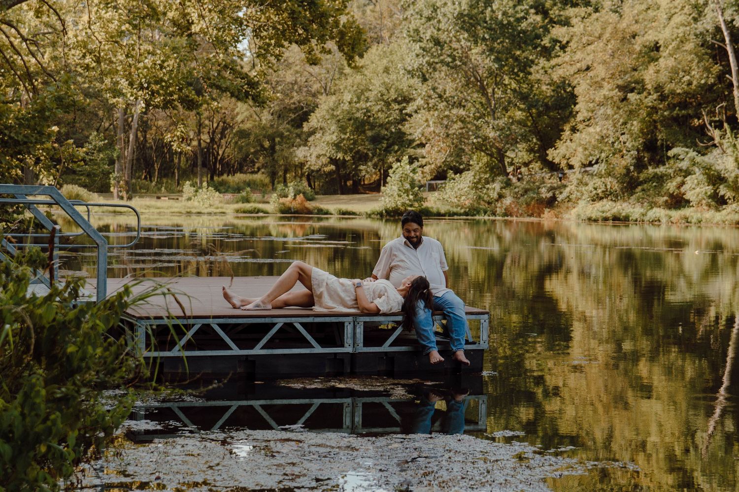 Couple relaxing on a dock by a serene reflective lake surrounded by lush green trees in a romantic outdoor setting.