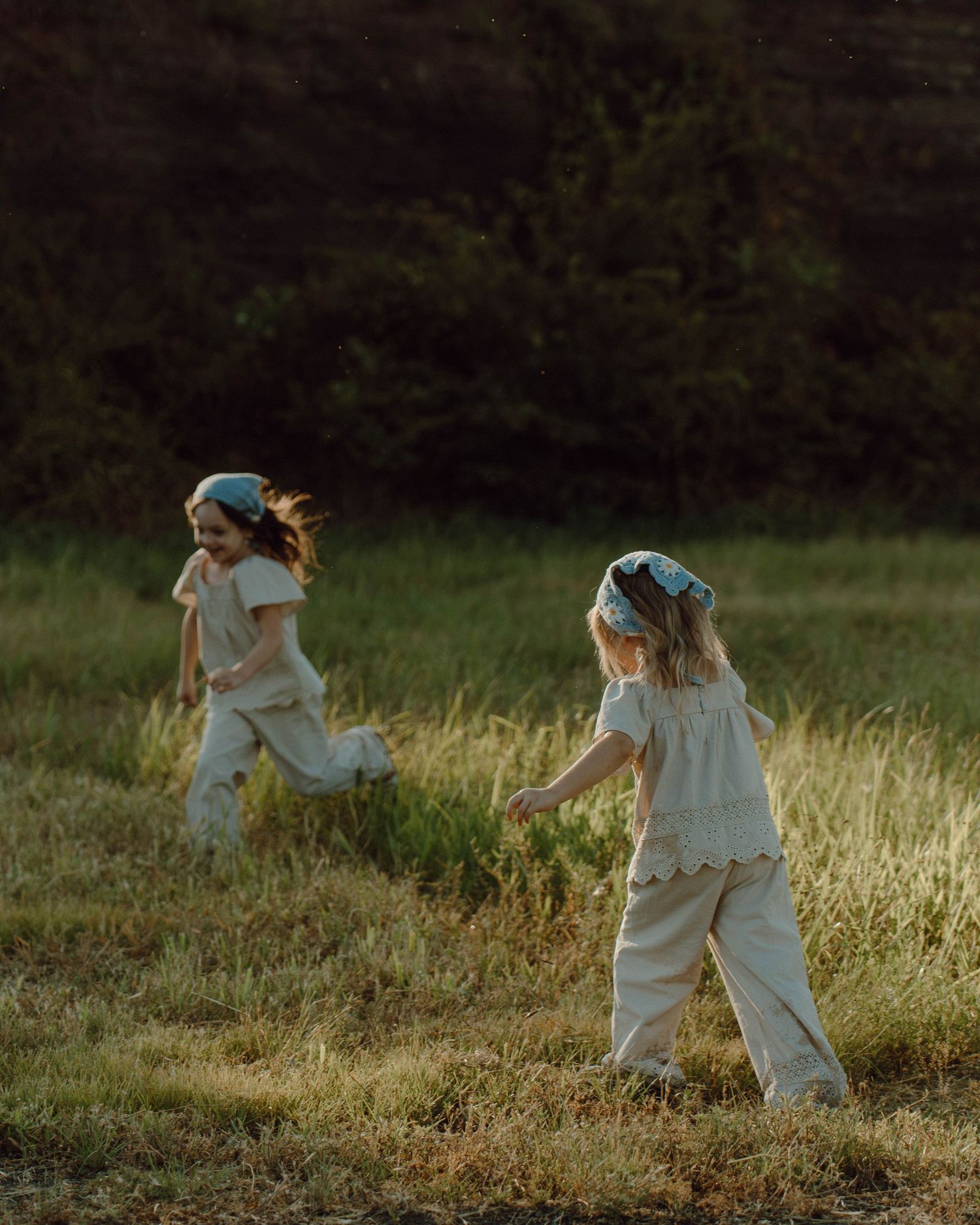 Two young girls in matching cream outfits and blue headbands running through a sunlit grassy field.