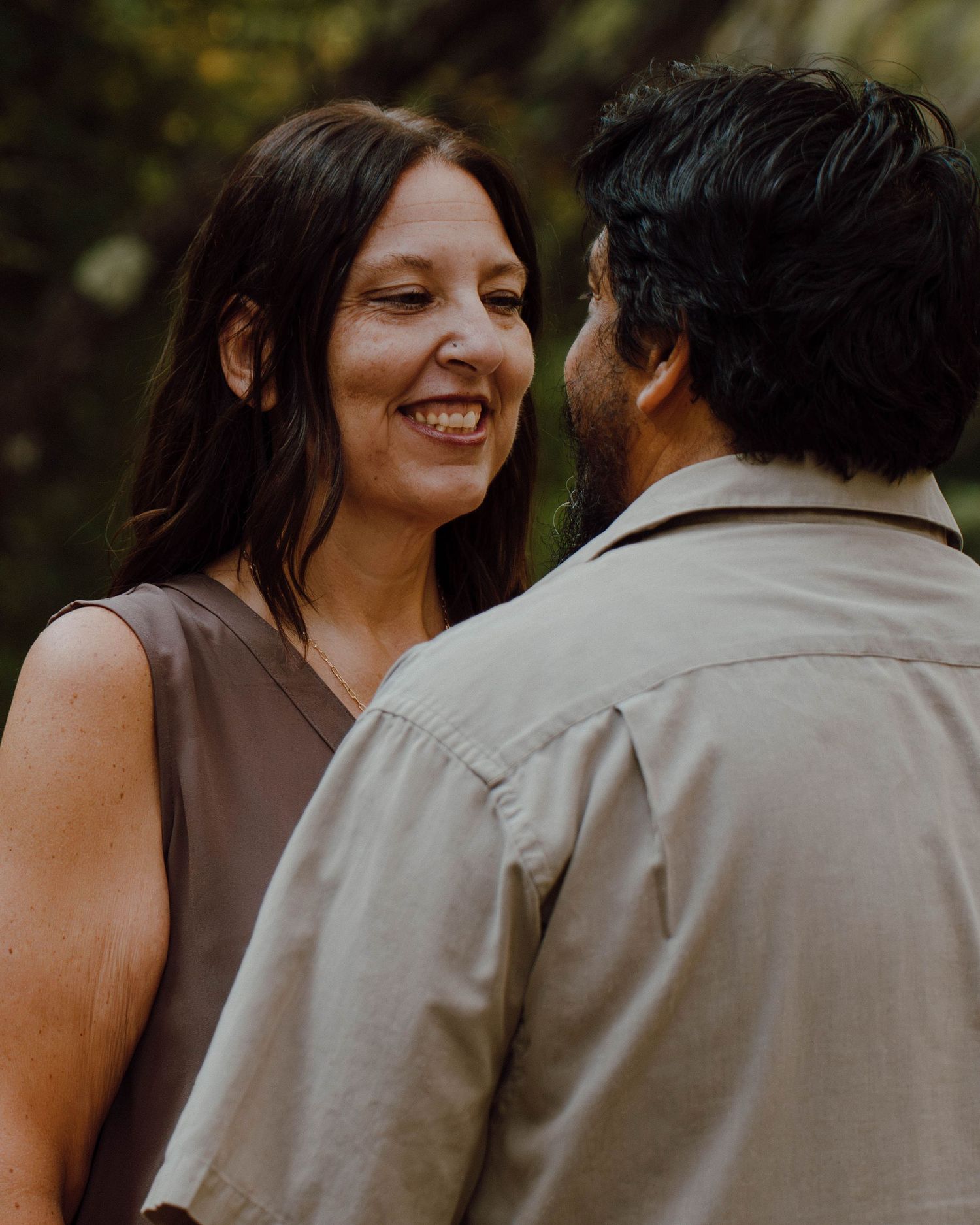 A smiling woman in a brown top gazes at a man in a beige shirt outdoors in a lush green setting.
