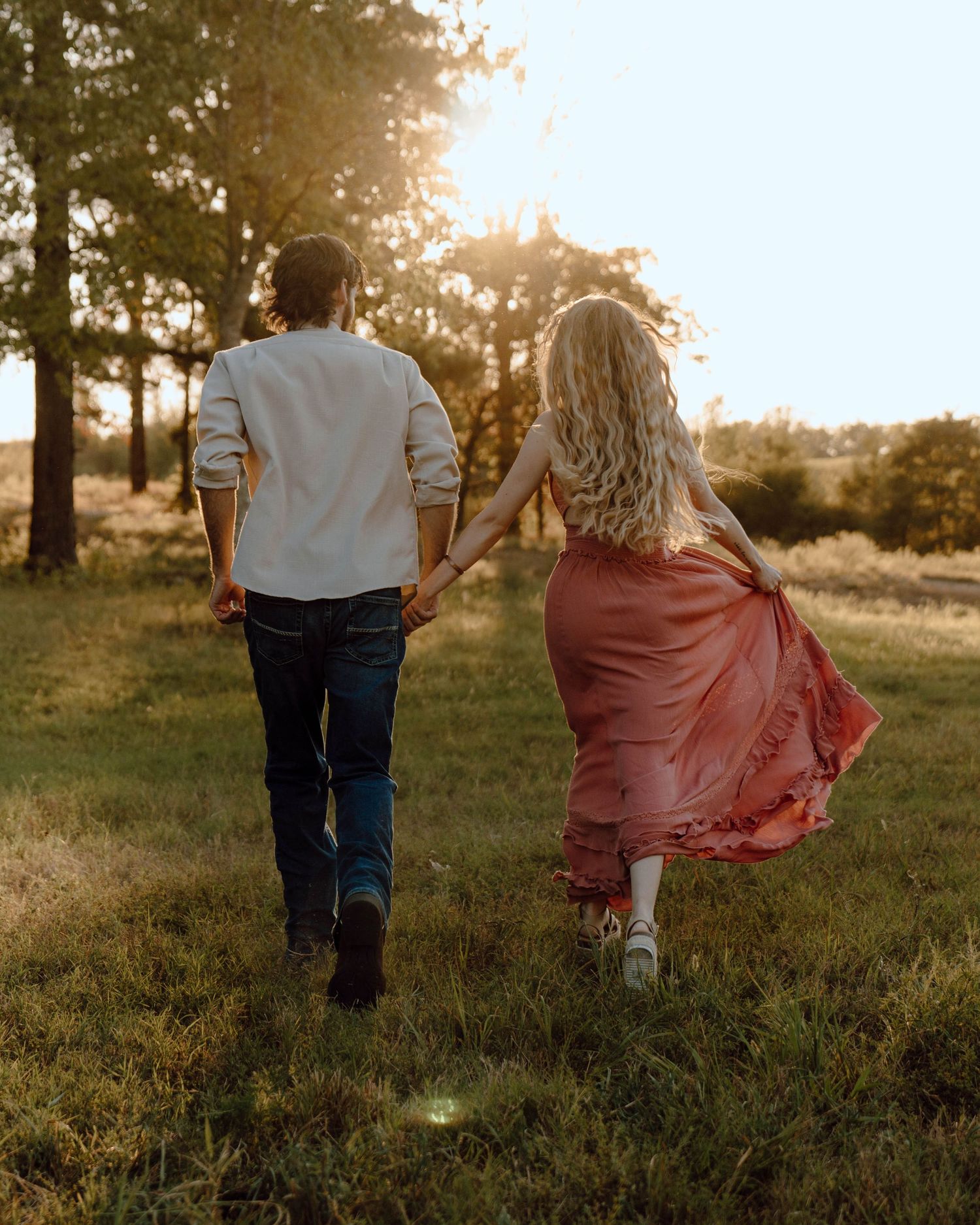Couple holding hands walking through sunlit field at golden hour, woman in flowing pink dress, man in white shirt and jeans.