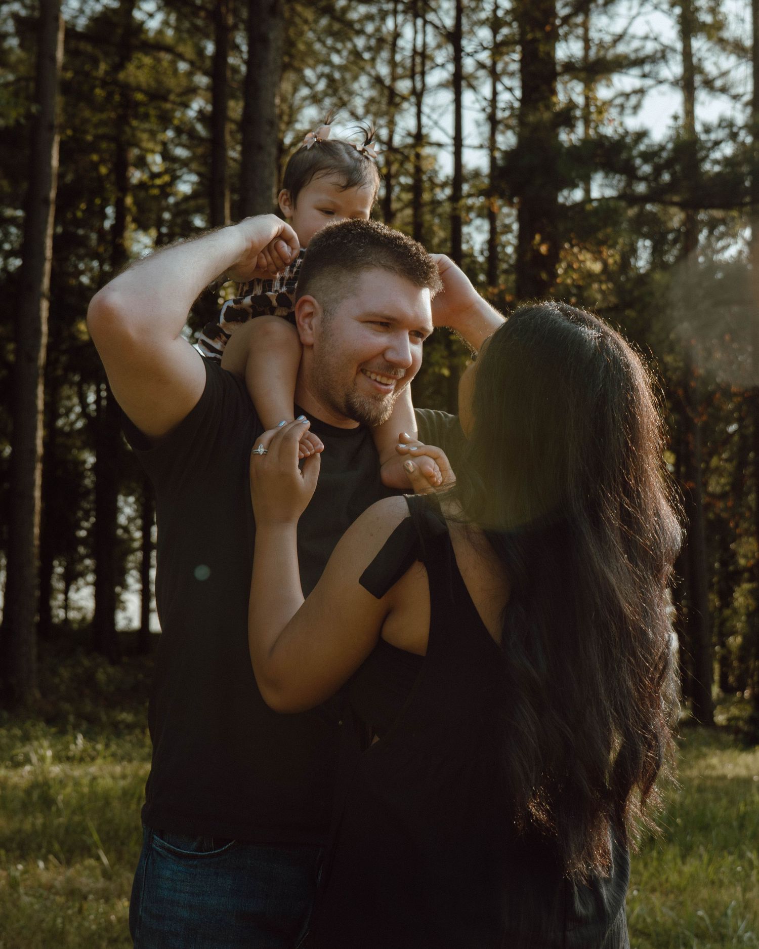 A happy family with a toddler on dad's shoulders sharing a joyful moment outdoors in a sunlit forest.
