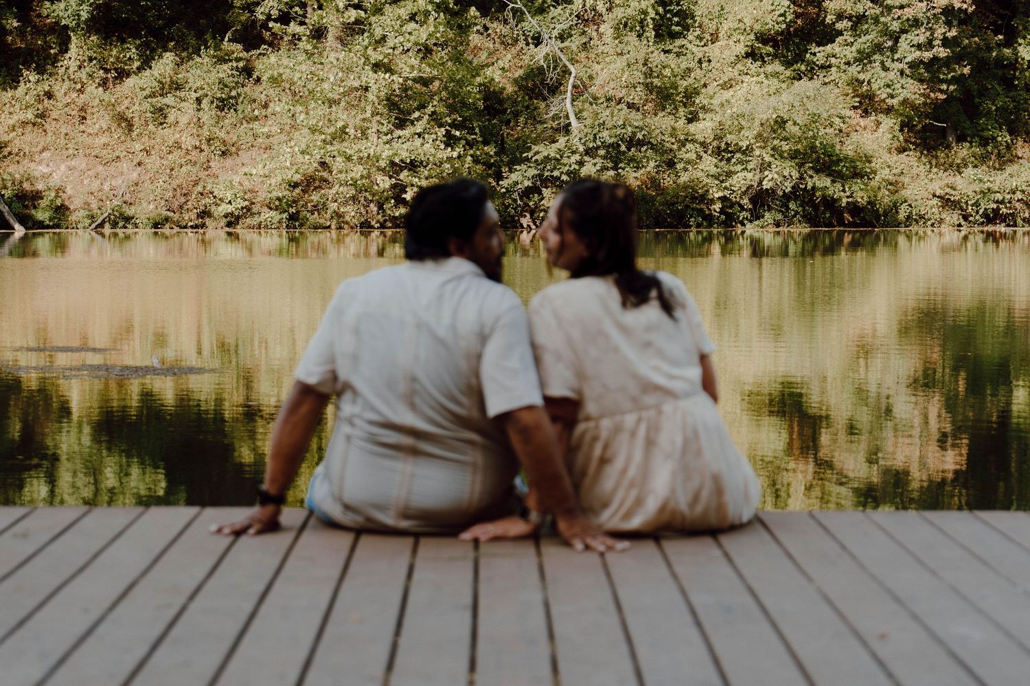 Couple sitting on a wooden dock by a tranquil lake, sharing a romantic moment surrounded by lush green trees.