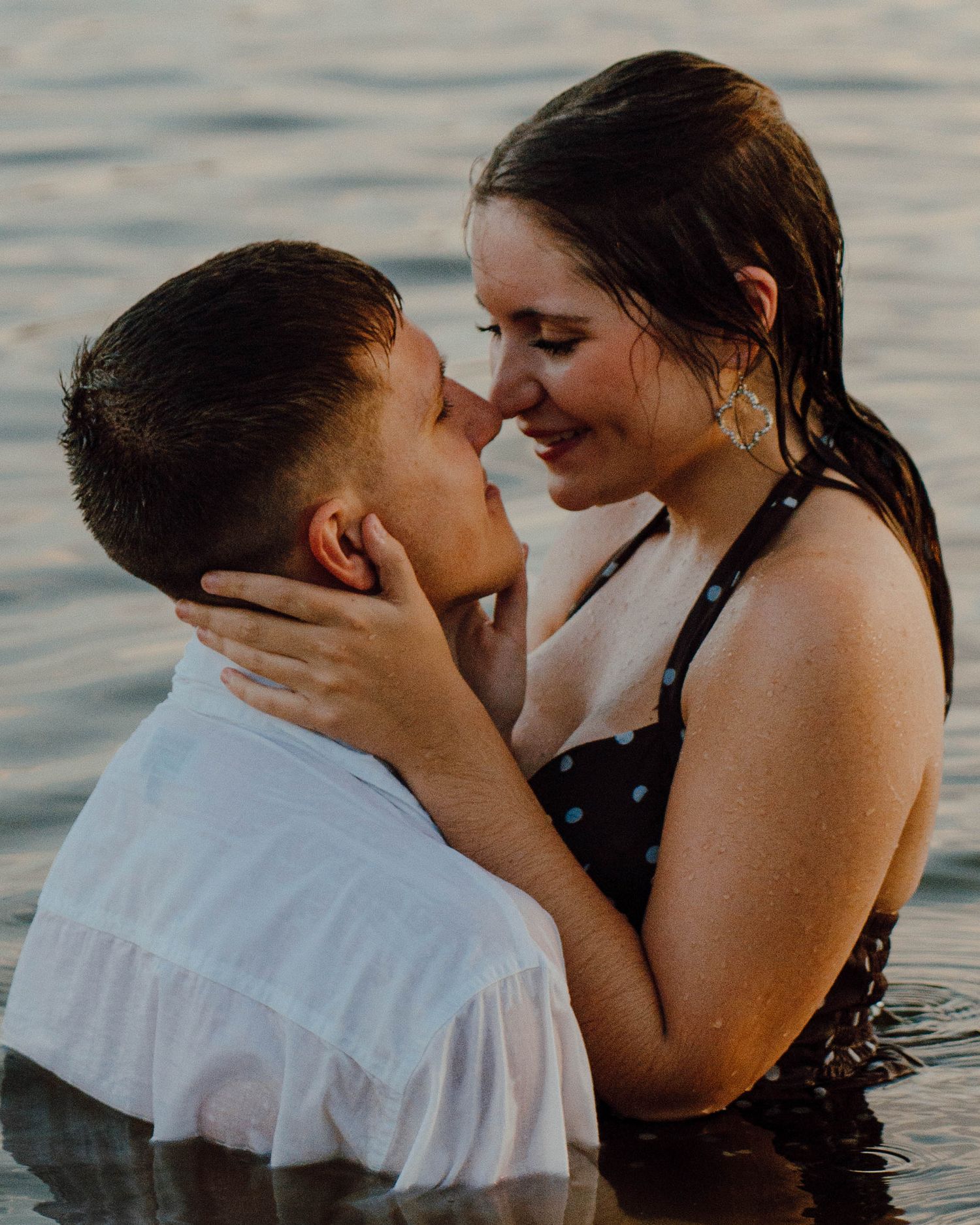 Couple sharing a romantic moment in water, smiling and embracing, wearing wet clothes at golden hour.