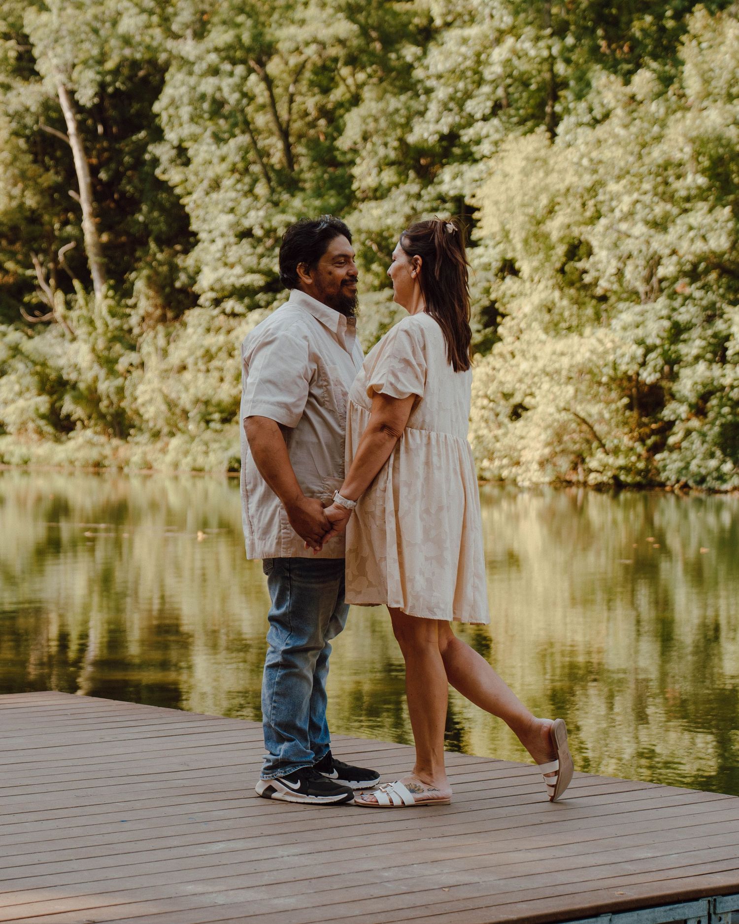 Couple holding hands on a wooden dock by a serene lake surrounded by lush green trees.