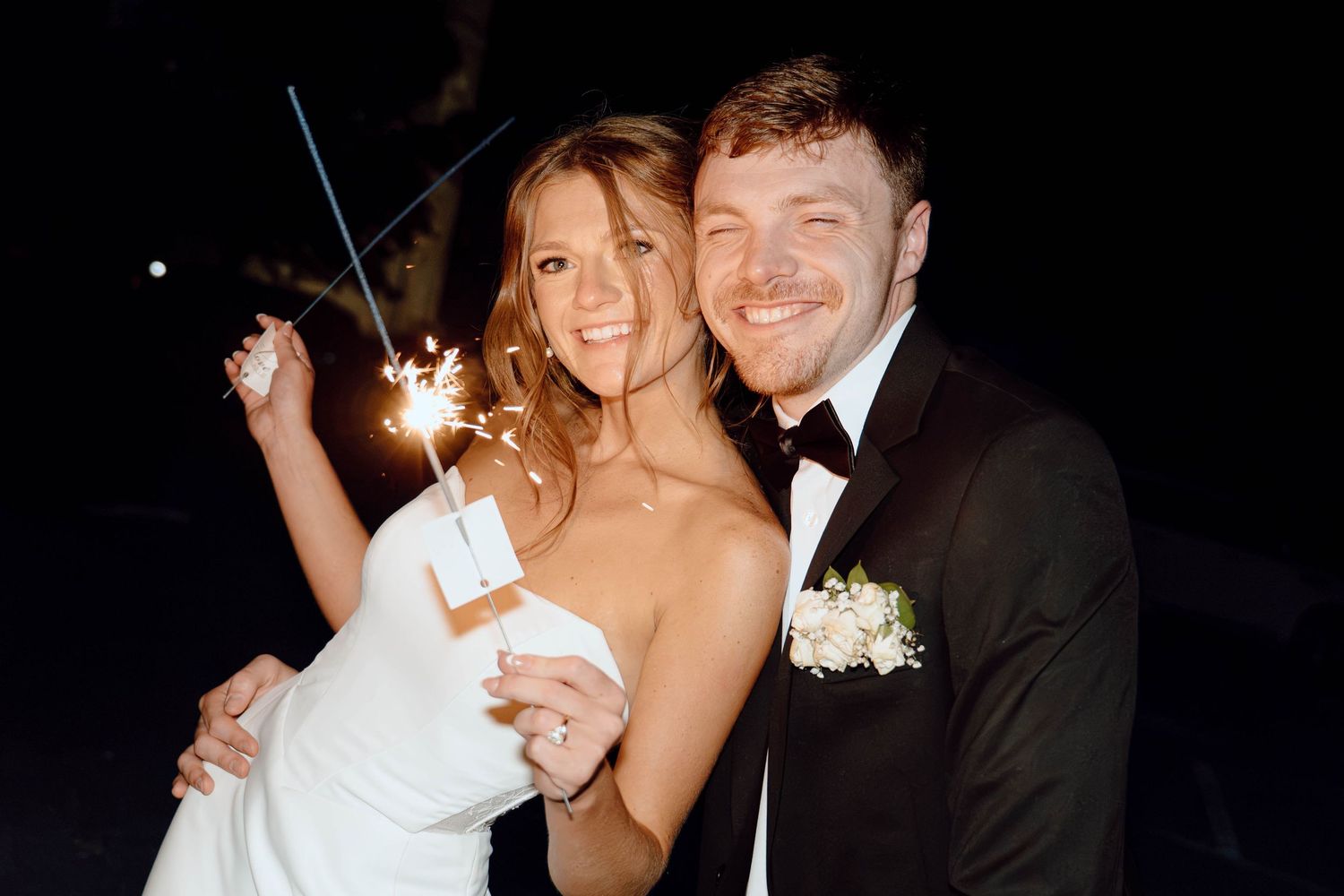 Smiling newlywed couple in wedding attire holding sparklers at night, groom in tuxedo and bride in white gown.
