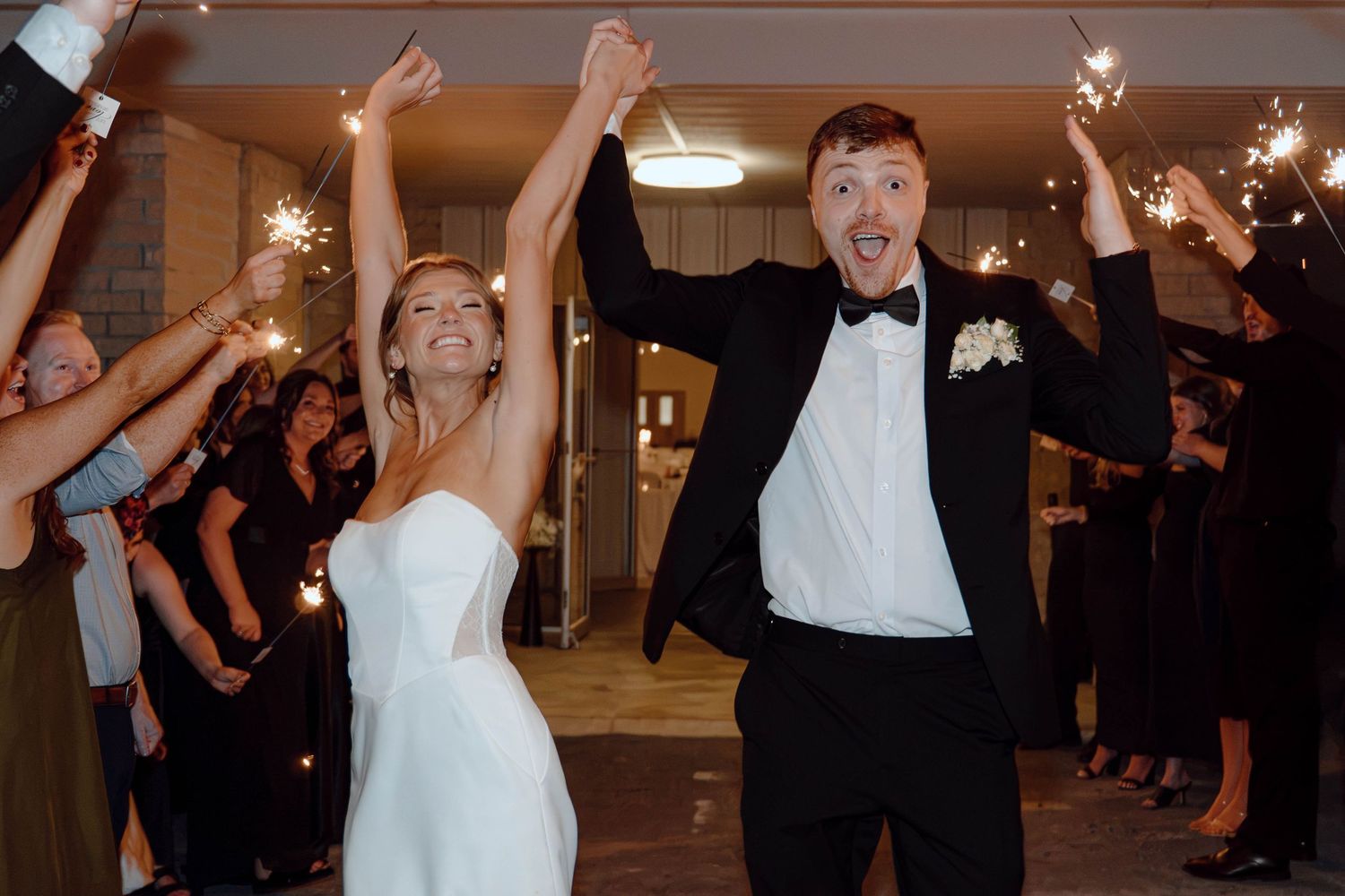 Joyful newlyweds celebrate with raised hands during sparkler send-off at elegant wedding reception.
