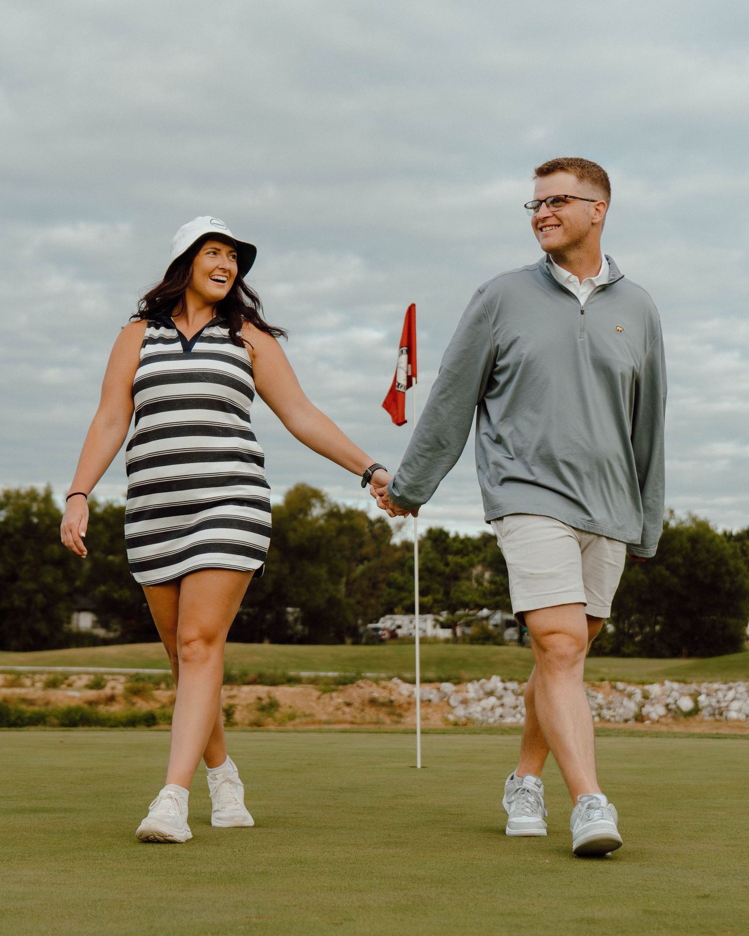 Happy couple holding hands walking on golf course green near flag in casual attire.