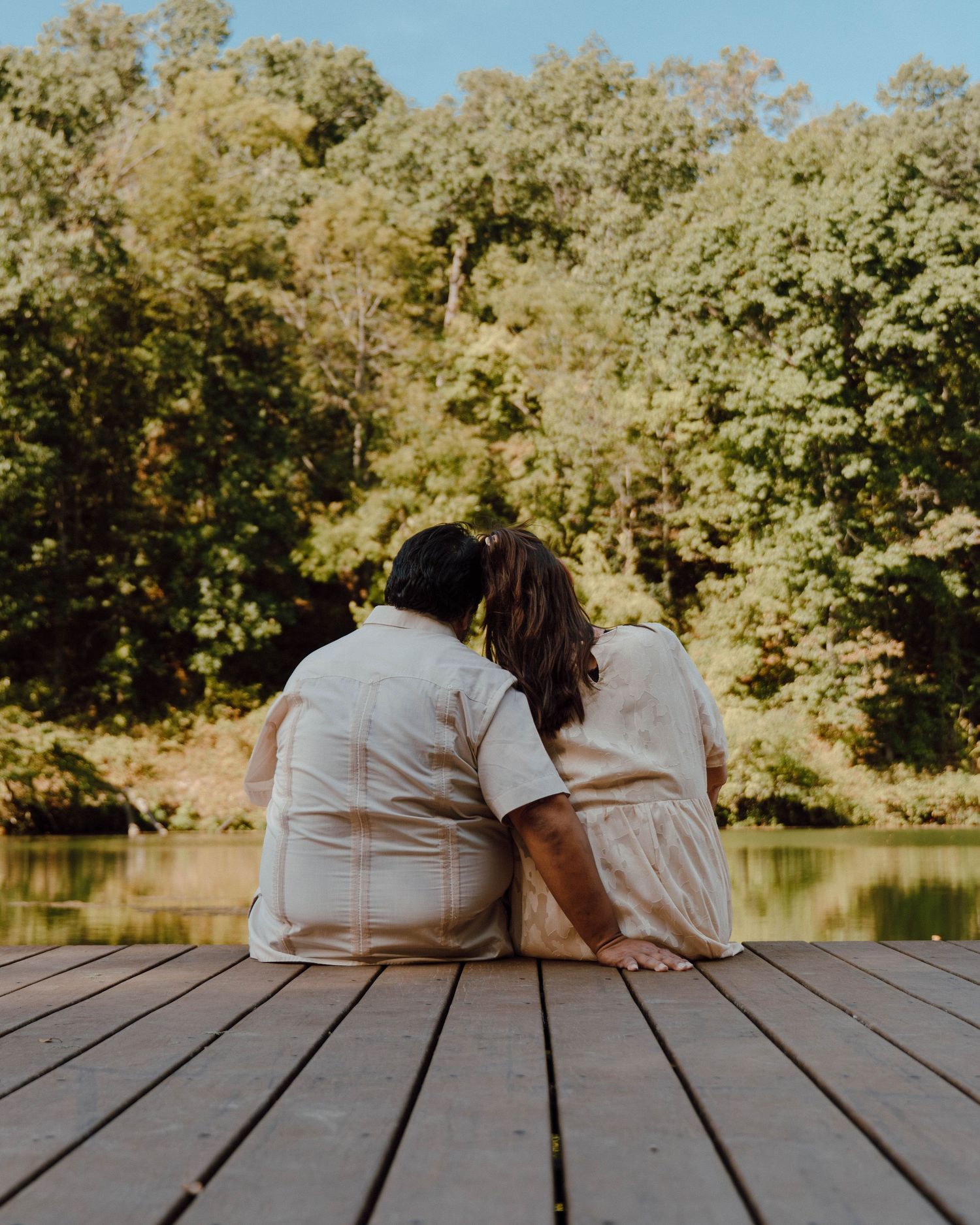 Couple sitting together on a wooden dock overlooking a serene lake surrounded by lush green trees.