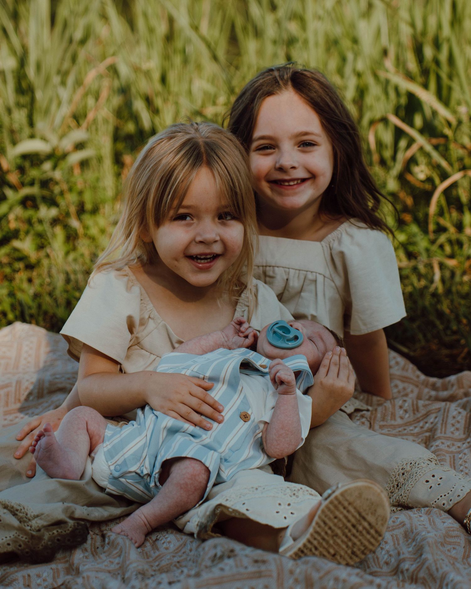 Two young girls in cream outfits sitting in a field holding a newborn baby on a blanket.