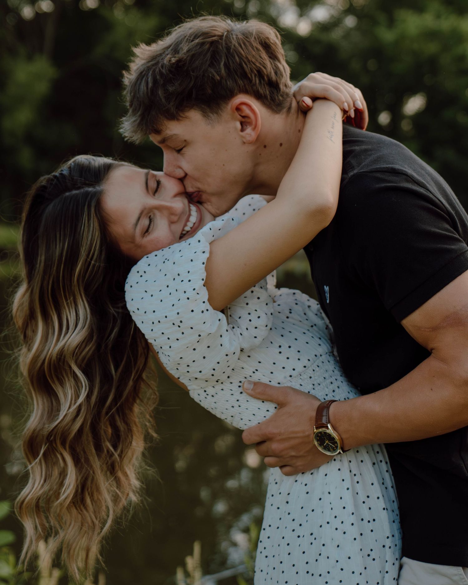 A young couple shares a romantic embrace outdoors, with the man kissing the smiling woman on the cheek near a serene pond.