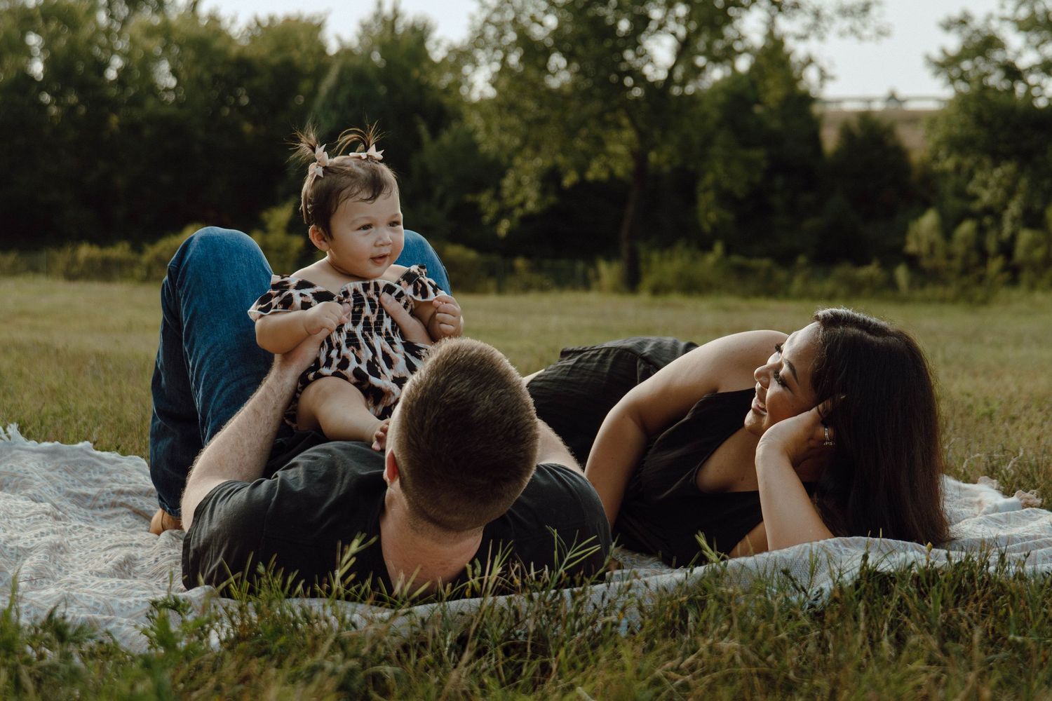 Young family lying on blanket in green field, parents playing with smiling baby in leopard print romper.