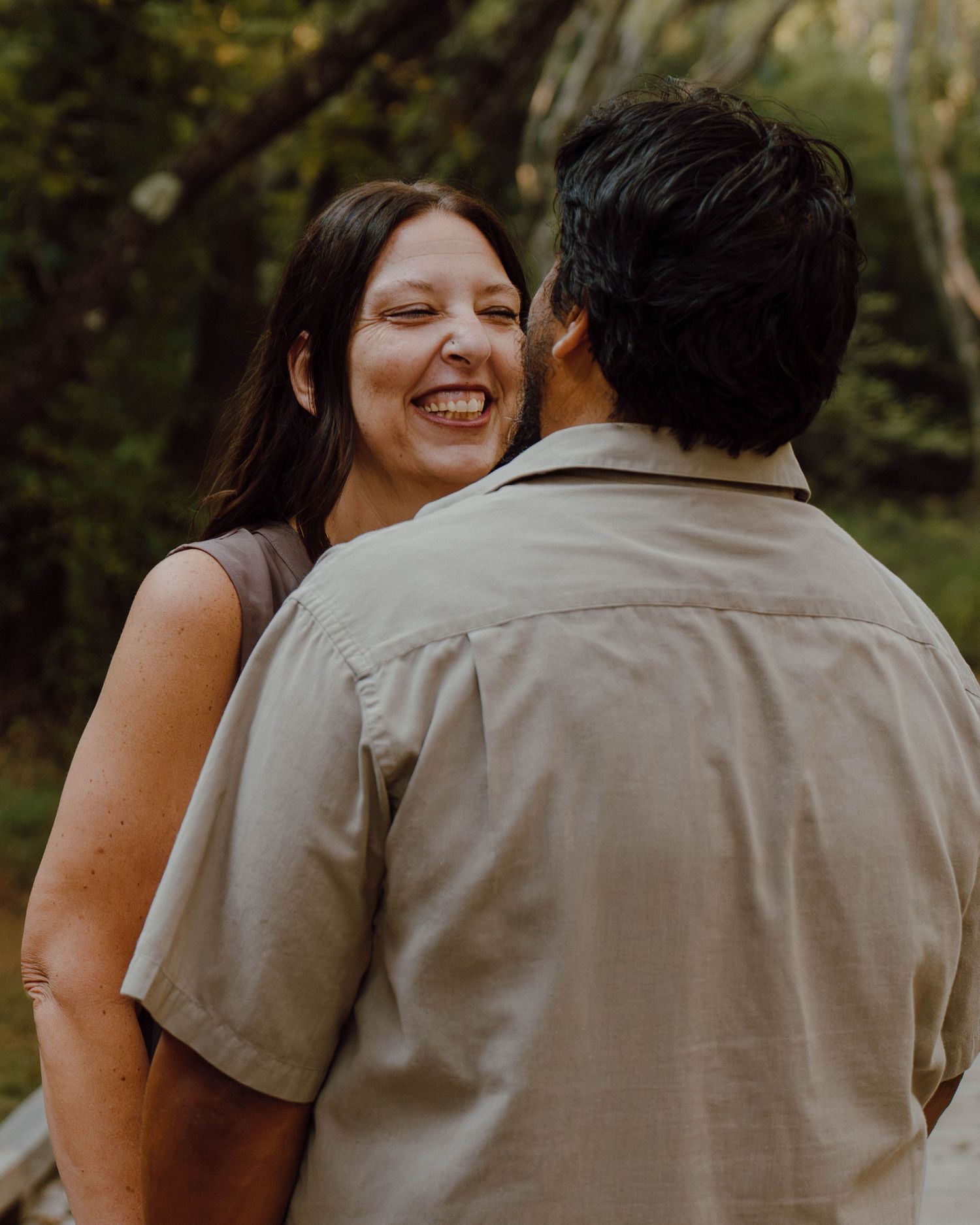 A laughing woman with long brown hair shares a joyful moment with a man in a beige shirt outdoors.