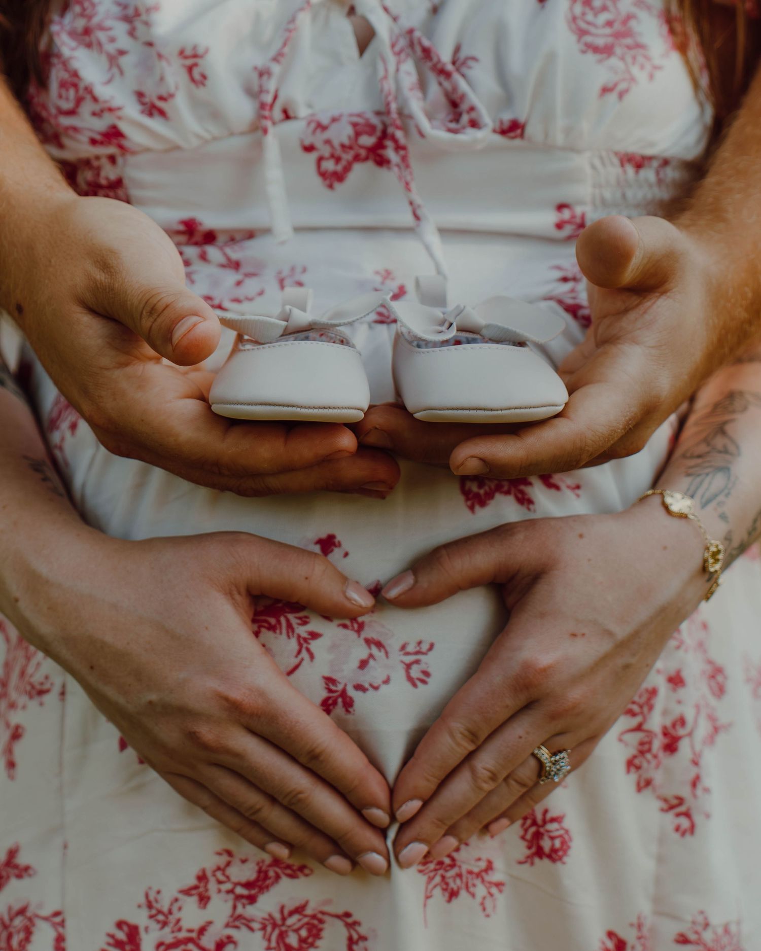 Couple forming heart shape with hands on pregnant belly while holding tiny white baby shoes.