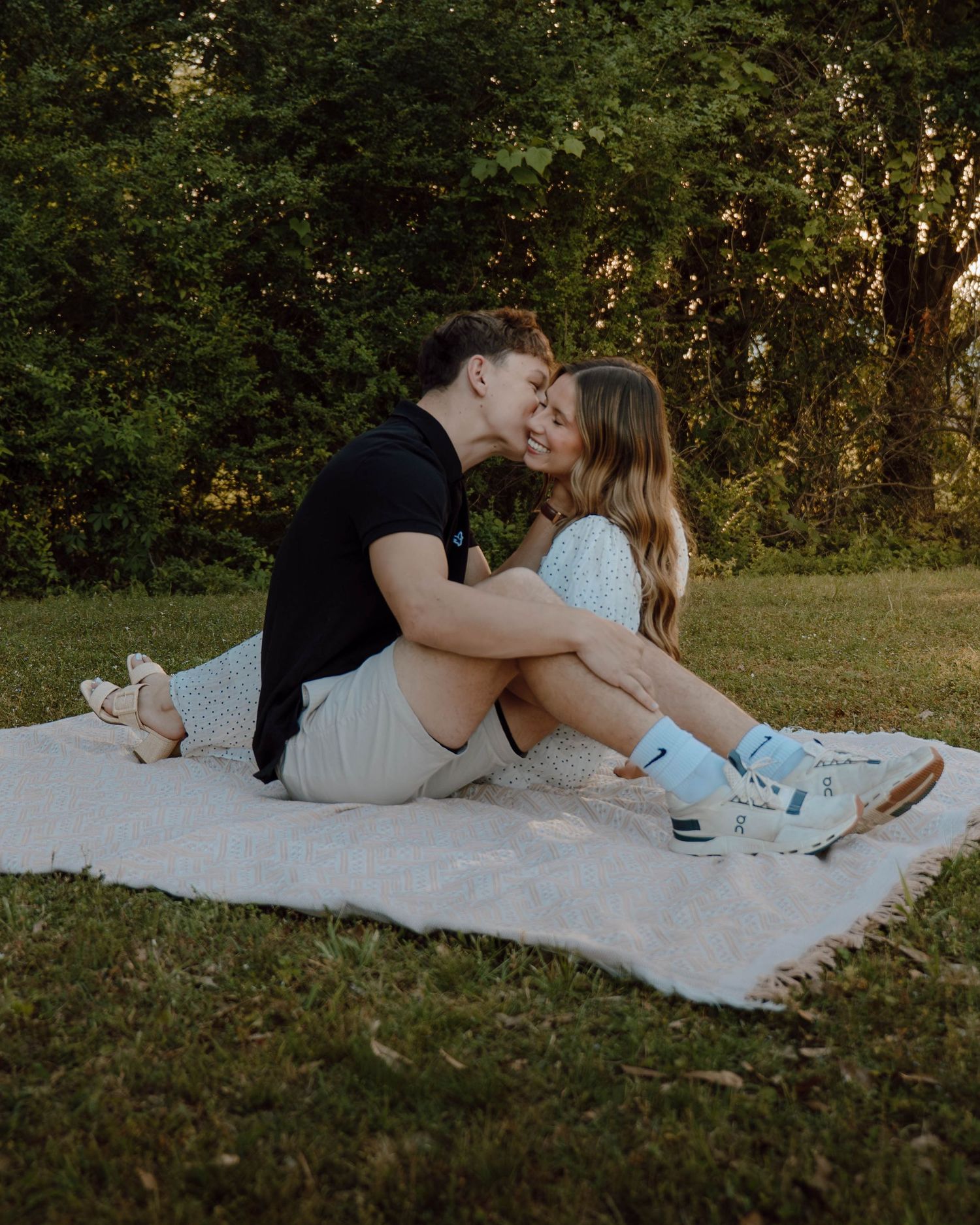 Couple sharing a romantic kiss while sitting on a blanket in a lush green park during golden hour.