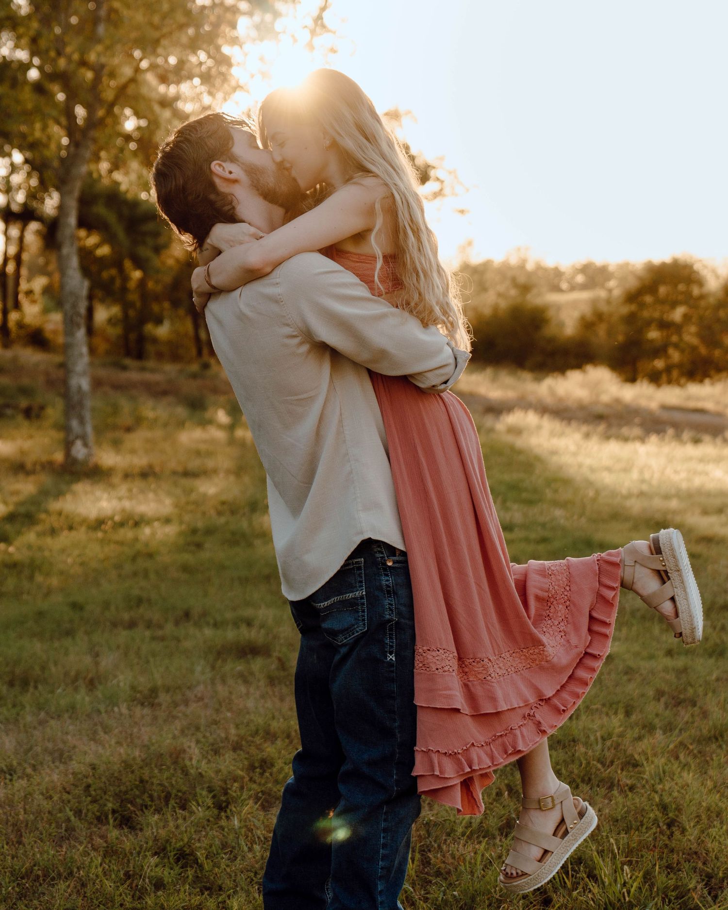 Couple sharing a romantic kiss at golden hour in a sunlit field, woman lifted off ground in pink dress.