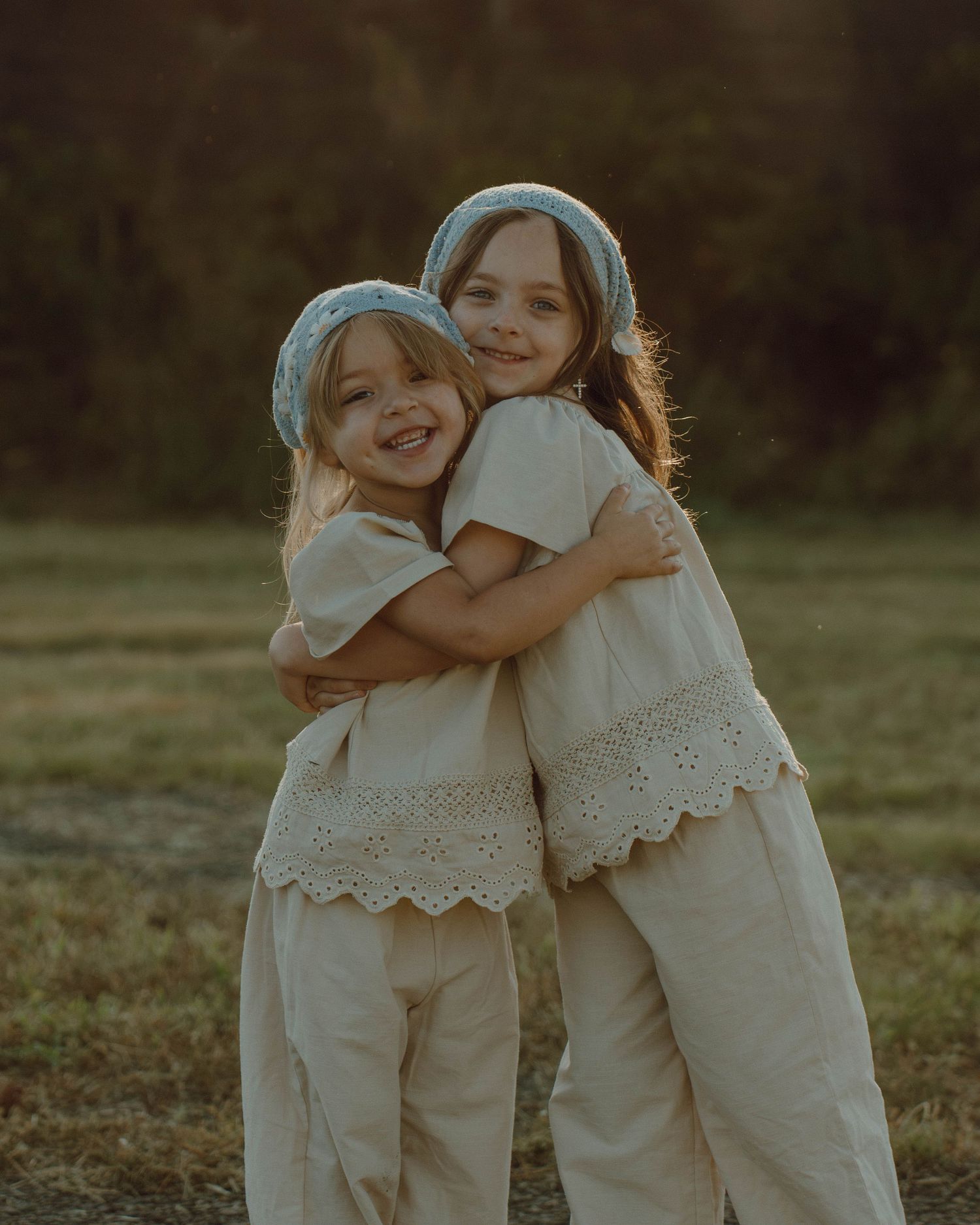 Two cheerful young sisters wearing matching cream outfits and blue headbands embracing in a sunny field.