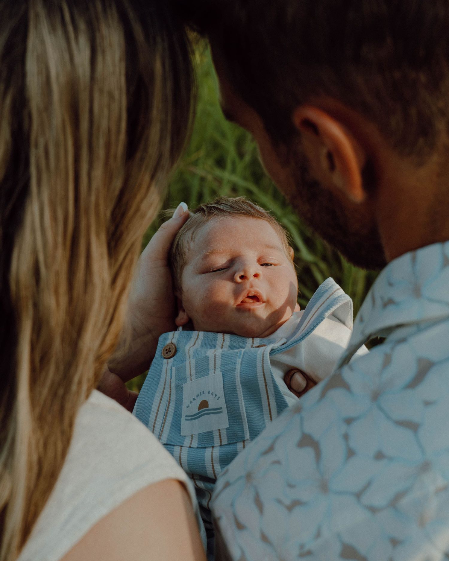 New parents tenderly cradling sleeping newborn baby in light blue outfit outdoors.