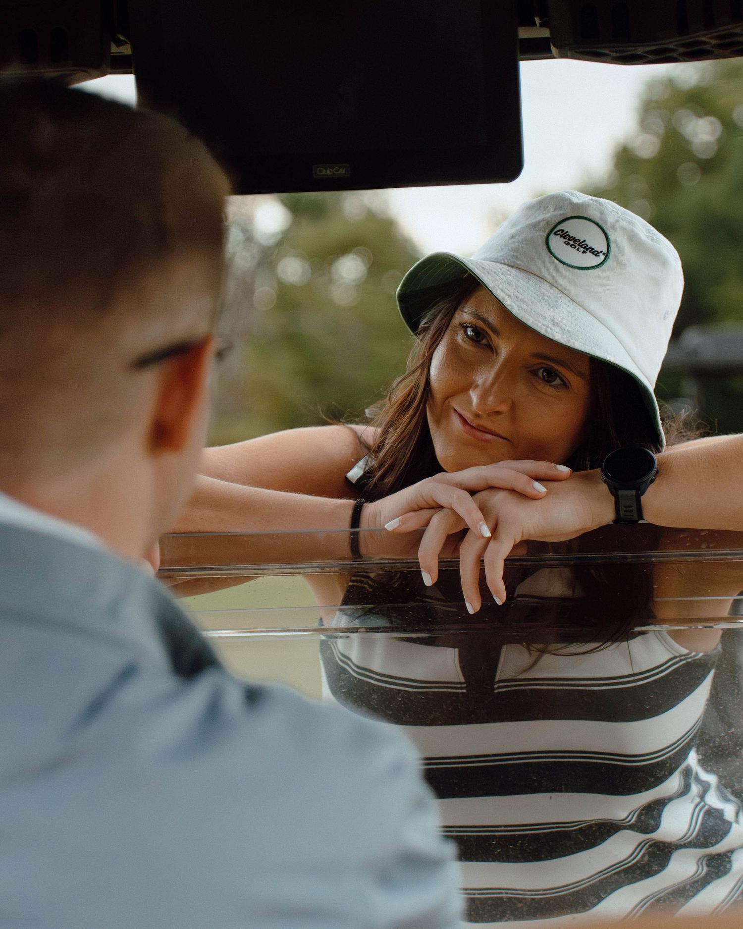 Woman in striped shirt and white bucket hat leans on car roof, gazing at person in foreground outdoors.