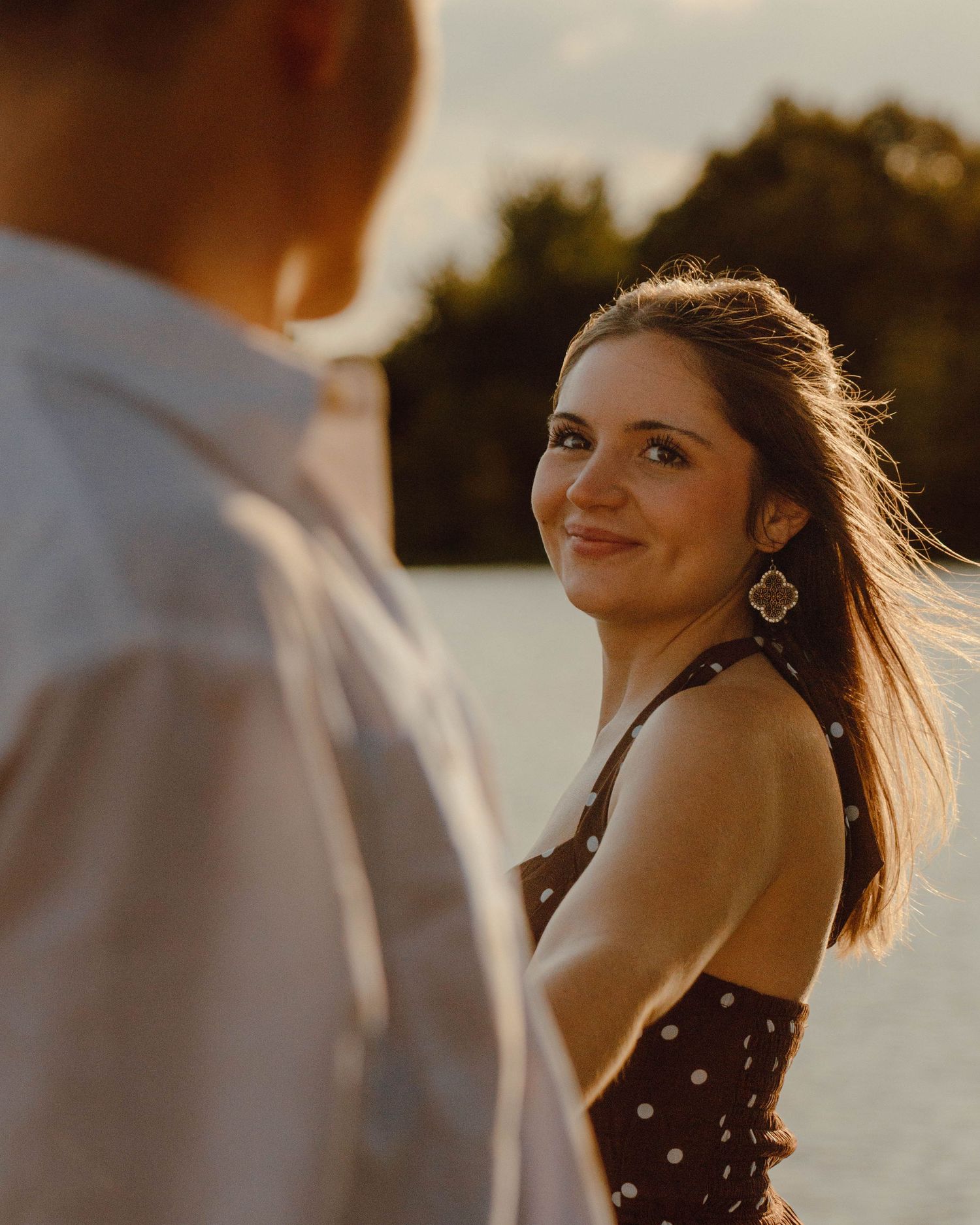 A smiling brunette woman in a polka dot dress glances back during a golden hour outdoor photo session near water.