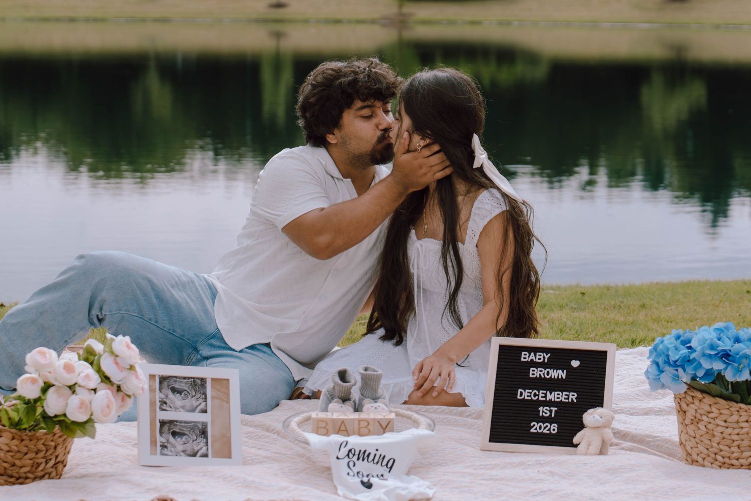 A couple kissing at an outdoor picnic by a lake with a baby announcement letterboard sign nearby.