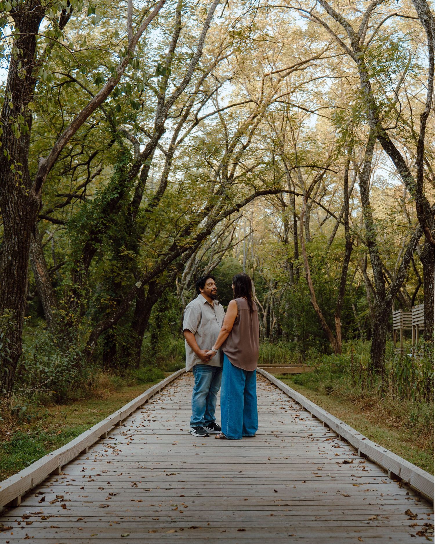 Couple standing face-to-face on a wooden boardwalk path surrounded by tall autumn trees in a lush forest setting.