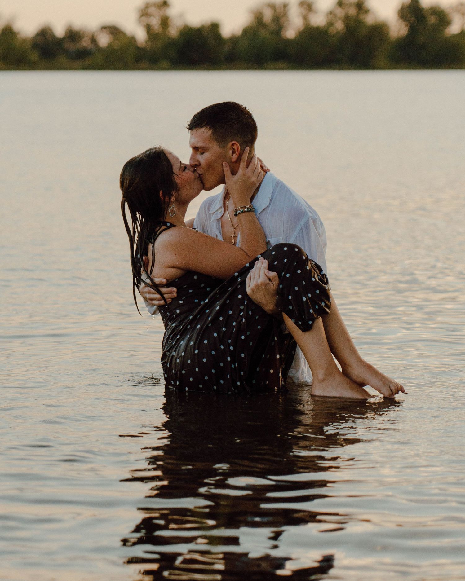 Couple sharing a romantic kiss while standing in a lake at sunset, with trees reflected in the calm water.