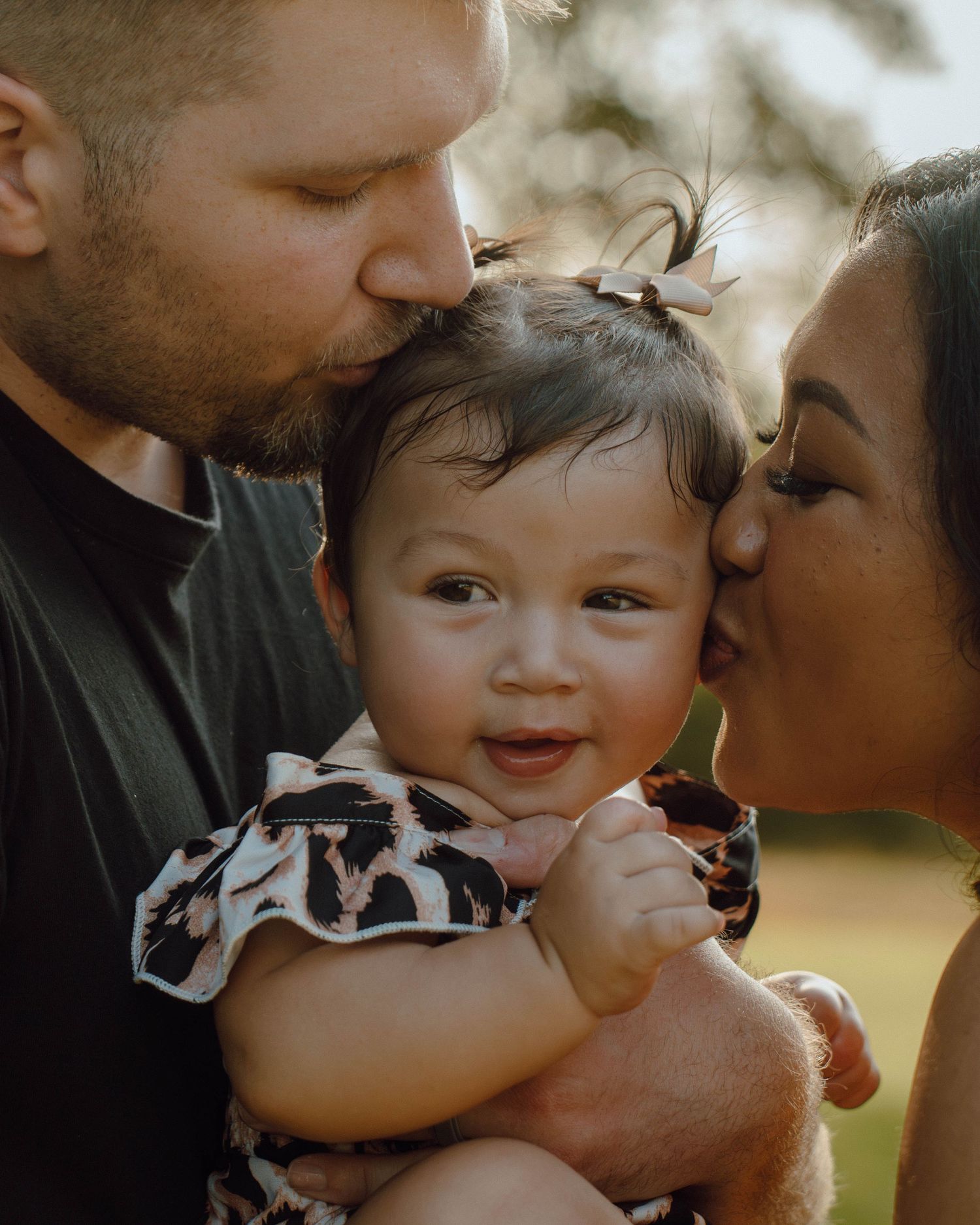 Parents kissing their smiling baby girl on cheeks in outdoor family portrait.