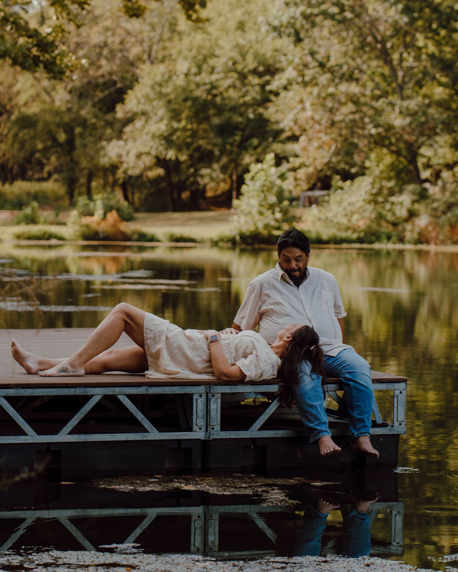 Couple relaxing on a wooden dock by a serene lake surrounded by lush green trees in a romantic outdoor setting.
