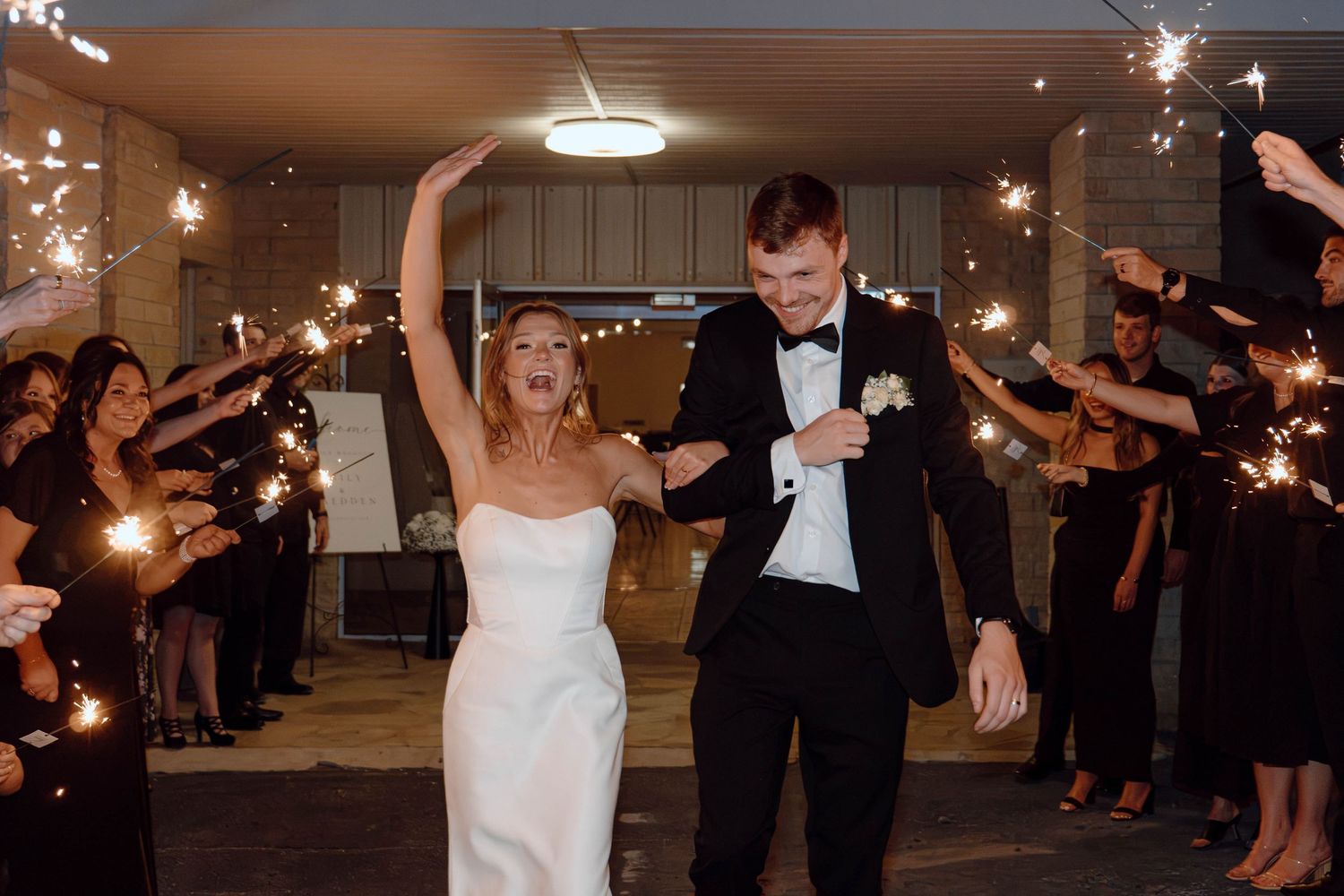 Bride and groom walk through sparkler exit tunnel at night, celebrating their wedding reception with cheering guests.
