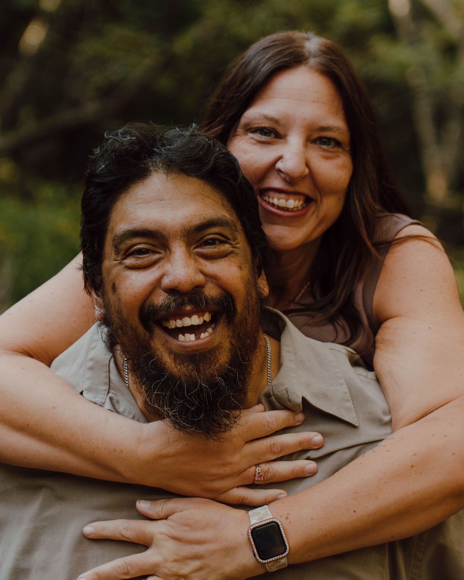 A bearded man and smiling woman share a joyful embrace outdoors, laughing together in a lush green setting.