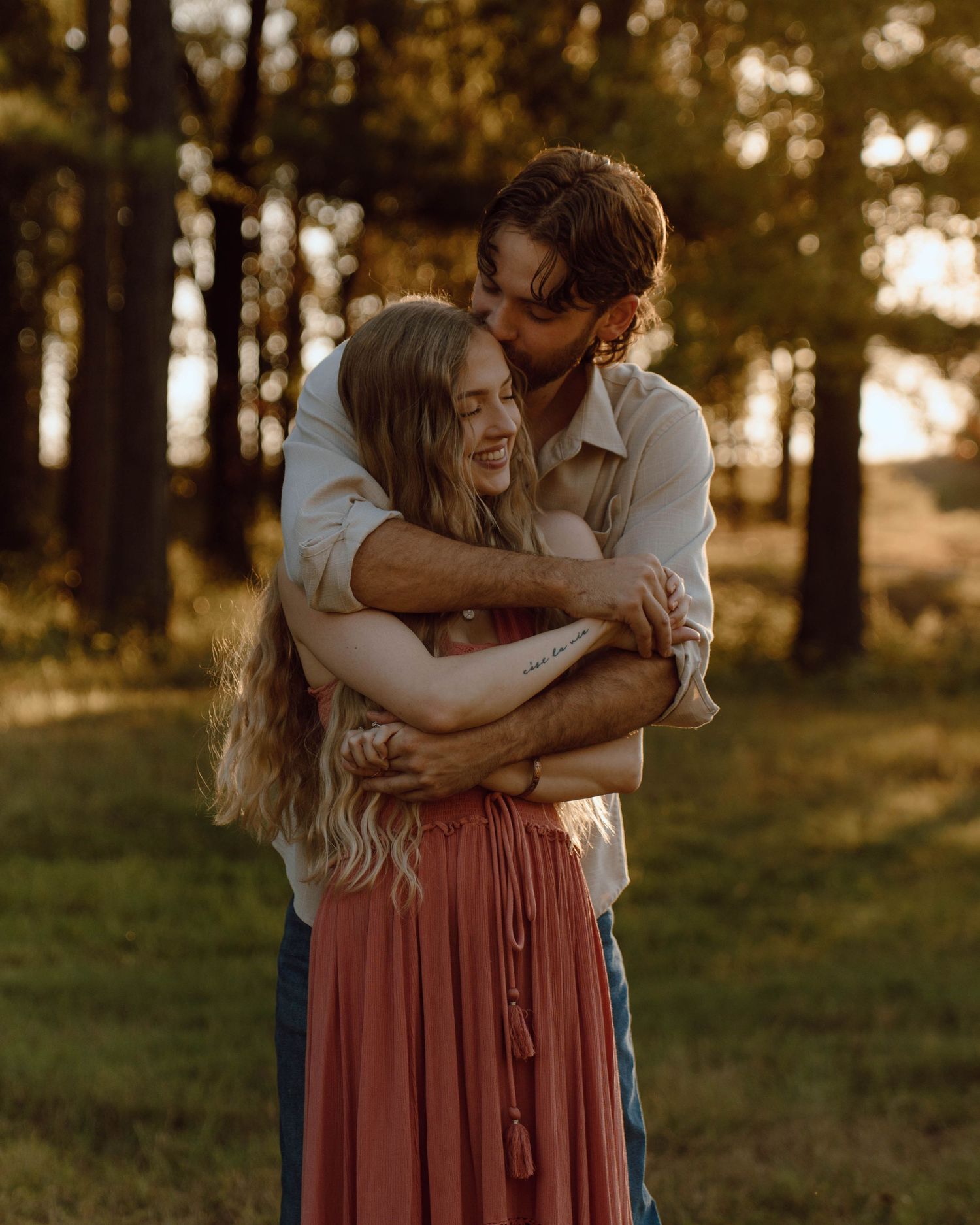 A couple embraces warmly in a sunlit forest, the man kissing the woman's head as she smiles in a pink maxi skirt.