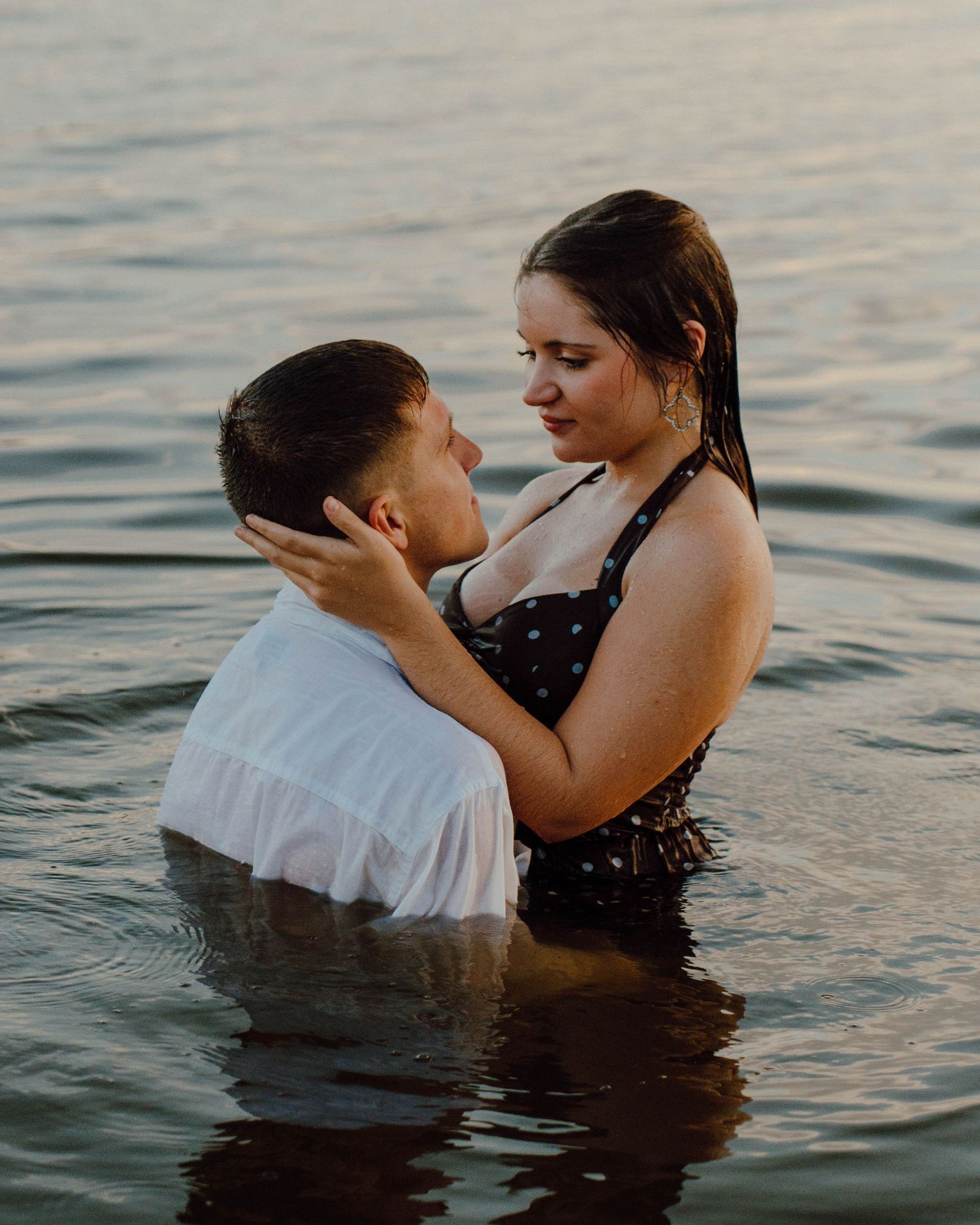 Couple embracing romantically in water at sunset, woman in polka dot swimsuit and man in white shirt.