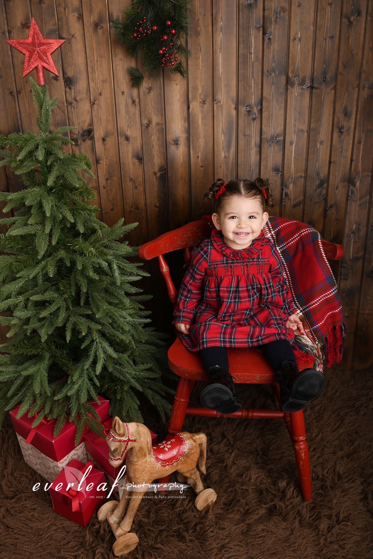 Young child in red Christmas clothes seated beside a rustic tree in the Everleaf Photography studio.