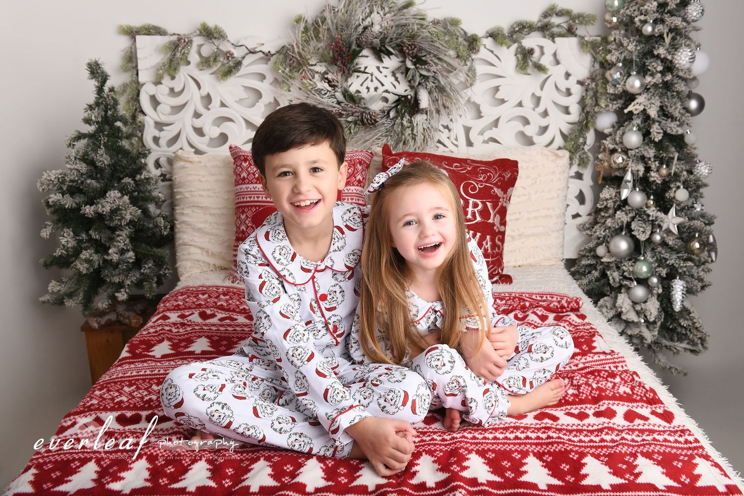 Two children in festive pyjamas on a red Christmas bed set at Everleaf Photography.