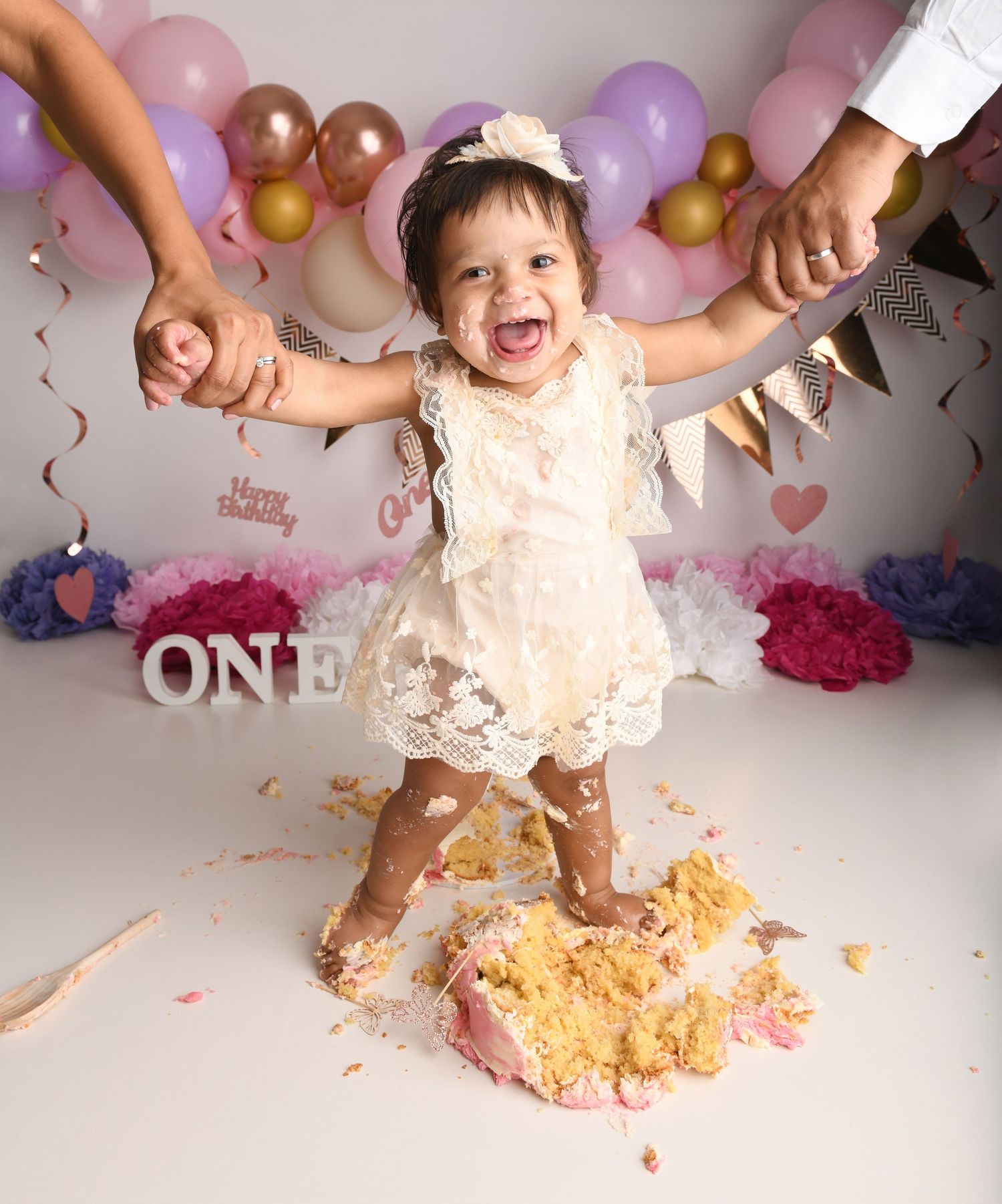 Baby enjoying a first birthday cake smash while holding their parents' hands