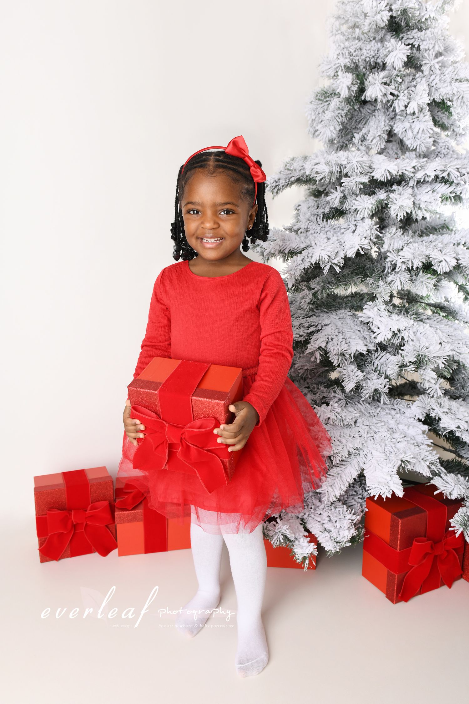 Young girl in a red Christmas dress beside a snow-covered tree and wrapped presents.