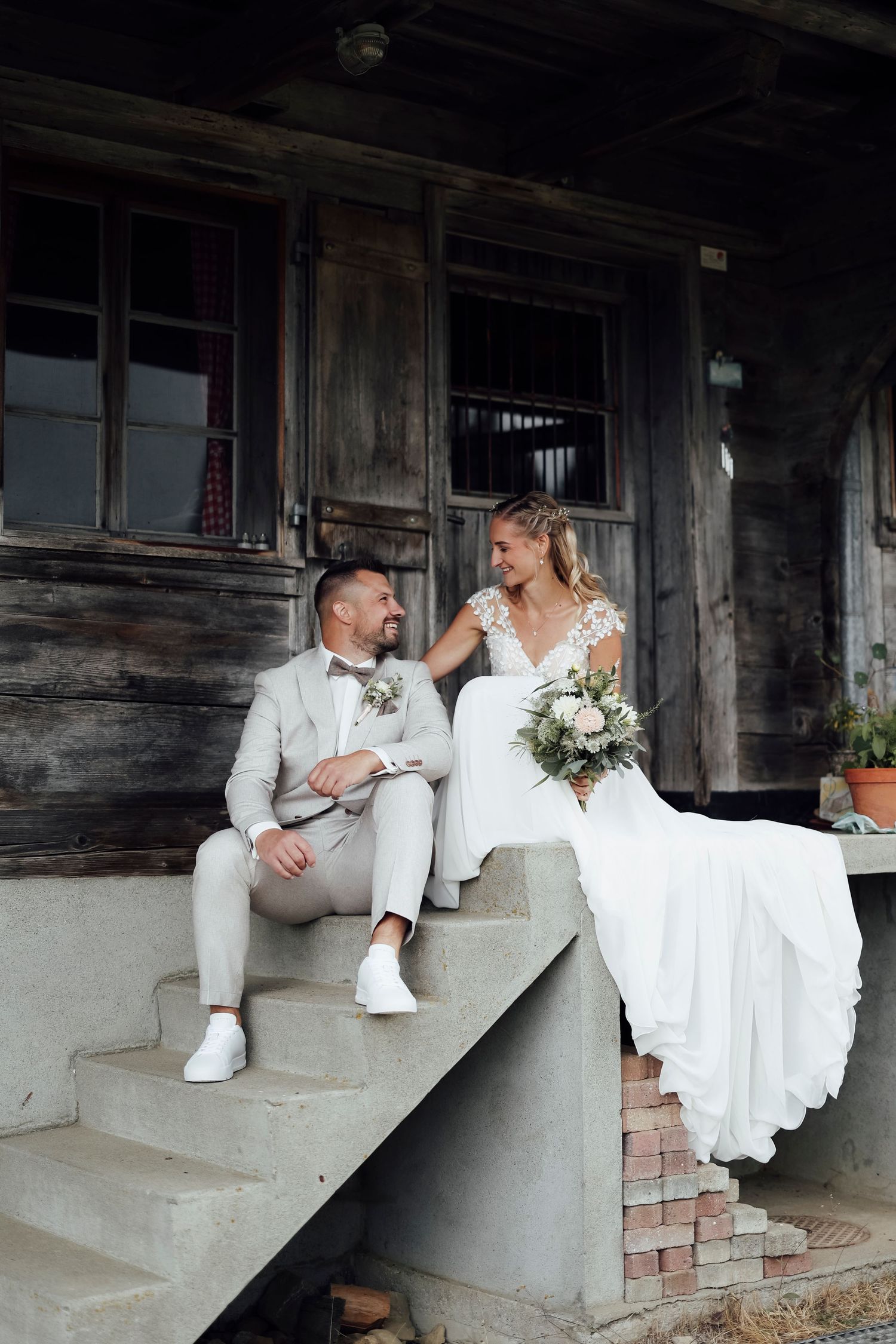 A smiling bride and groom sit on steps of a rustic wooden building on their wedding day.