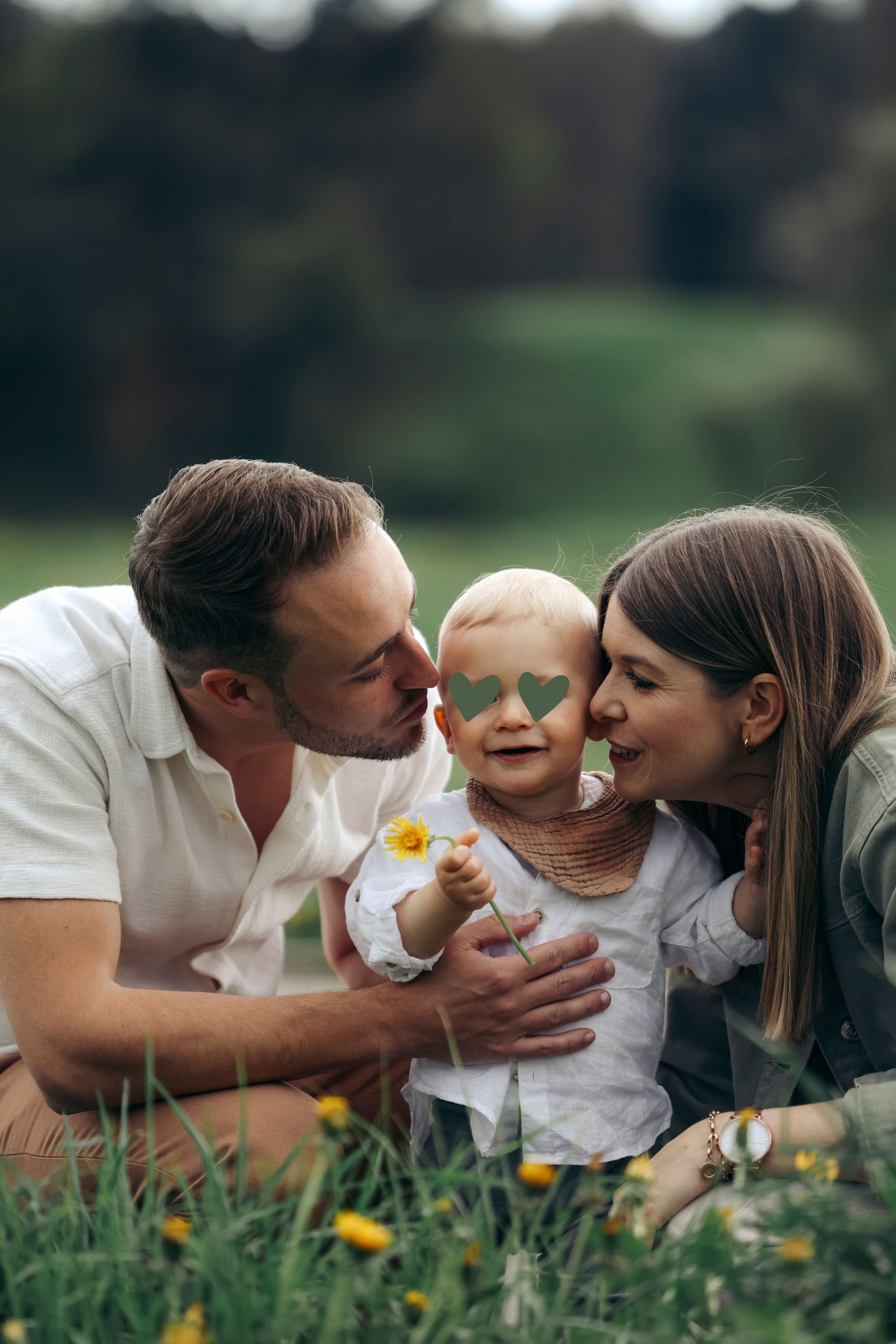 Happy parents kiss their baby on the cheeks while sitting in a flower-filled green meadow.
