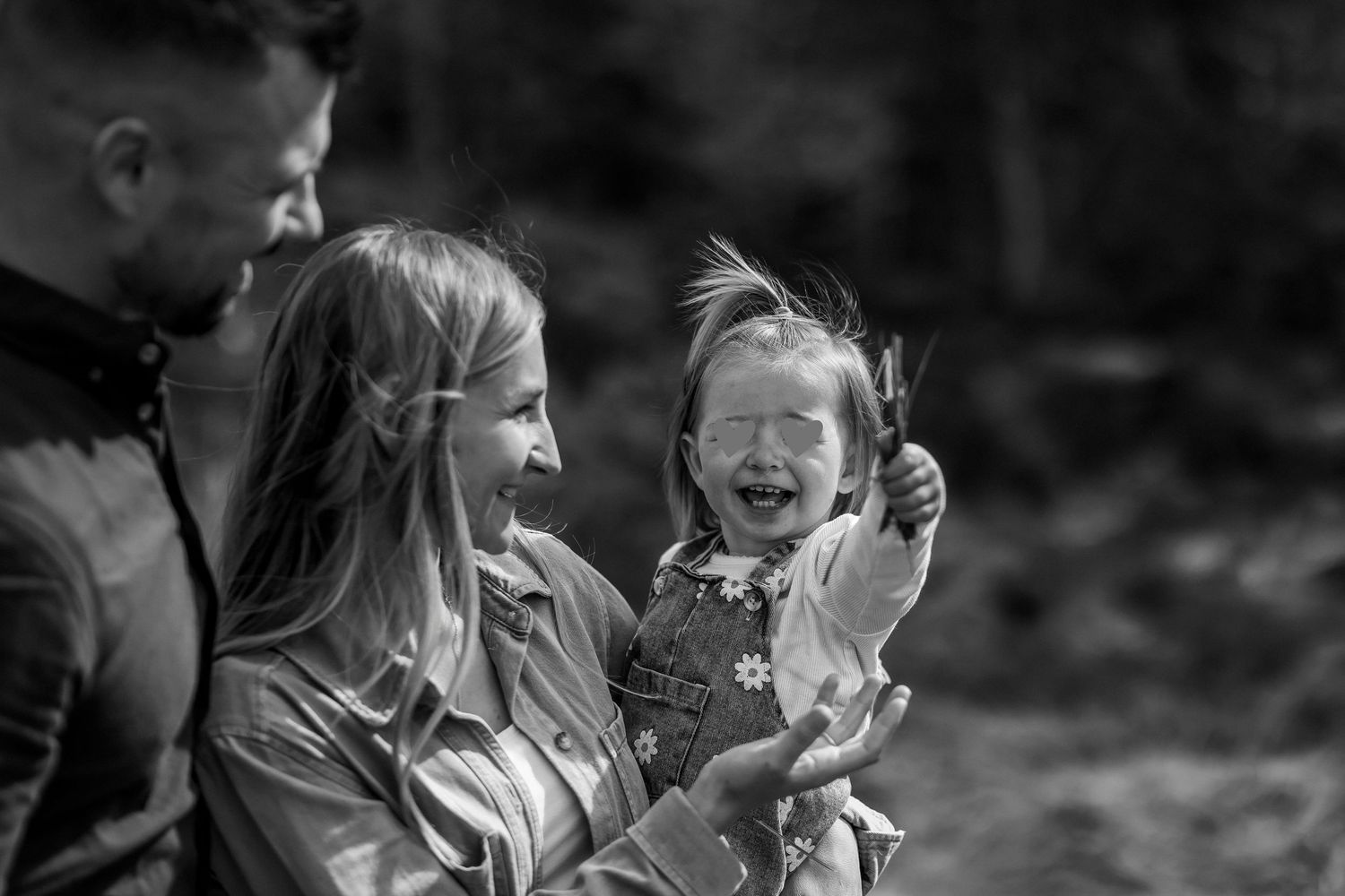 A laughing toddler is held by her smiling mother while father looks on in black and white photo.